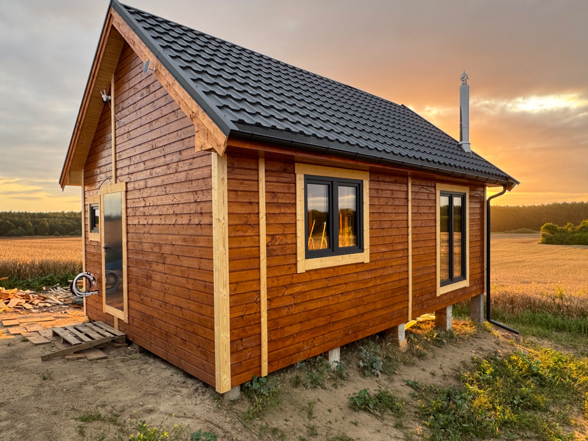 Gotowy, drewniany domek z czarnym dachem na tle zachodzącego słońca. Widoczne okna z czarnymi ramami, elementy wykończeniowe i fragment pola w tle.
