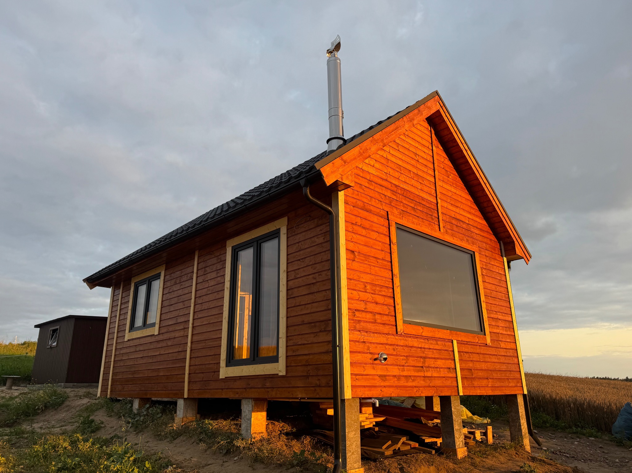 Drewniany, parterowy dom z kominem na fundamencie palowym, w ciepłym świetle zachodzącego słońca, z widocznym polem w tle. Widoczne czarne ramy okienne.