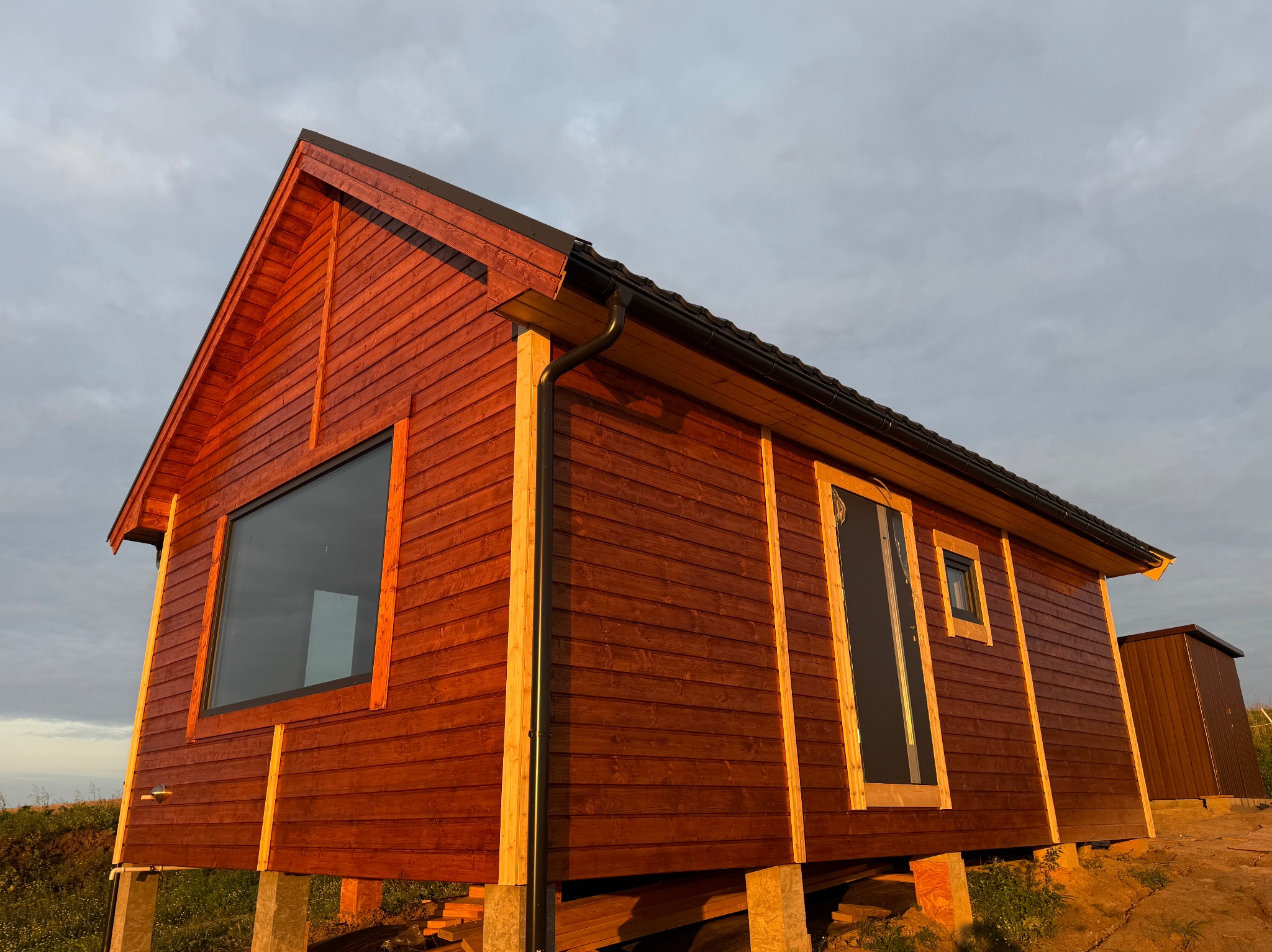 Czerwony dom szkieletowy z drewnianą elewacją, dużym oknem i czarną rynną, wzniesiony na betonowych podporach, na tle pochmurnego nieba.