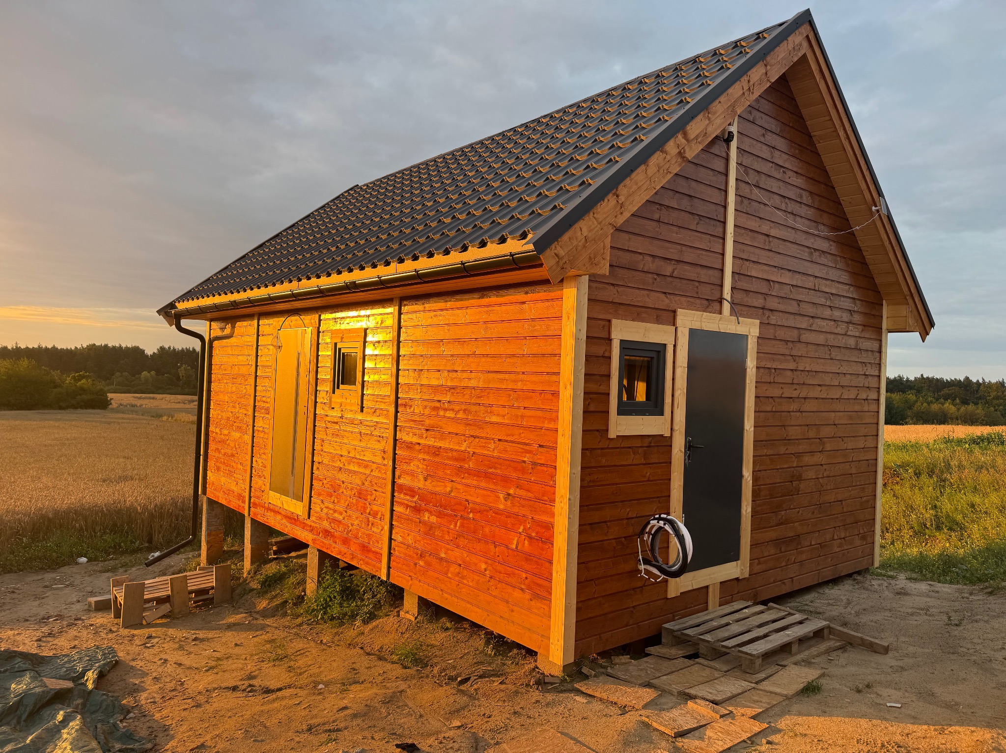 Drewniany dom z ciemnym dachem, oświetlony zachodzącym słońcem, z widocznymi detalami wykończenia i otaczającą go naturą.