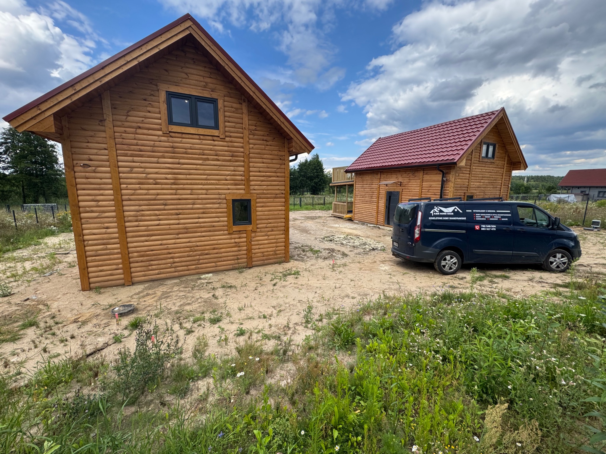 Dwa drewniane domy z bali, jeden z balkonem, na działce z samochodem dostawczym firmy budowlanej. Błękitne niebo z chmurami w tle.