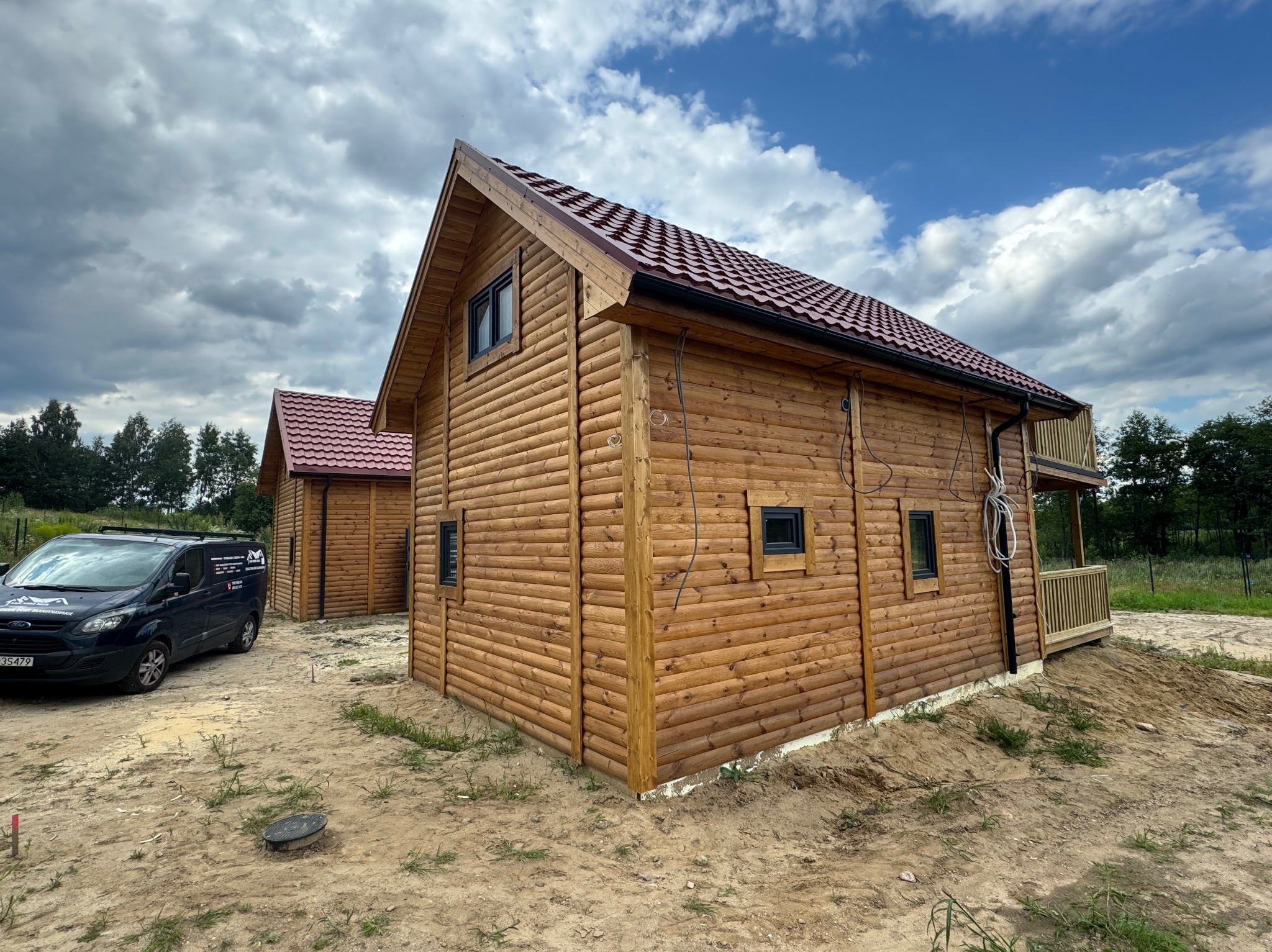 Dwa identyczne domki z bali z czerwonymi dachami, widoczne z zewnątrz, z niebieskim vanem z logo firmy w tle i częściowo odsłoniętą ziemią wokół.