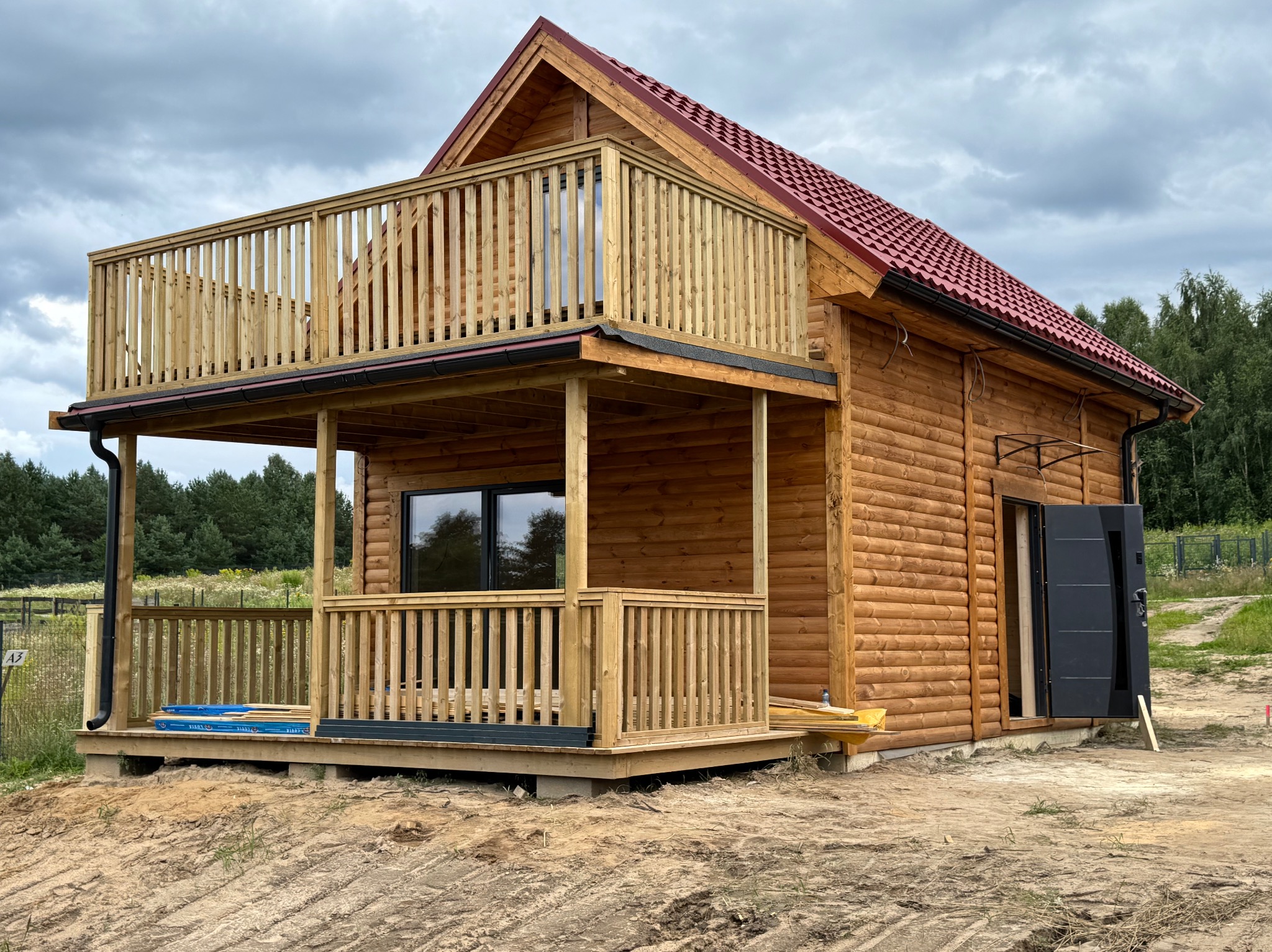 Dwukondygnacyjny, drewniany dom z balkonem i czerwonym dachem, w otoczeniu zieleni, na działce z piaskiem. Drzwi wejściowe otwarte.