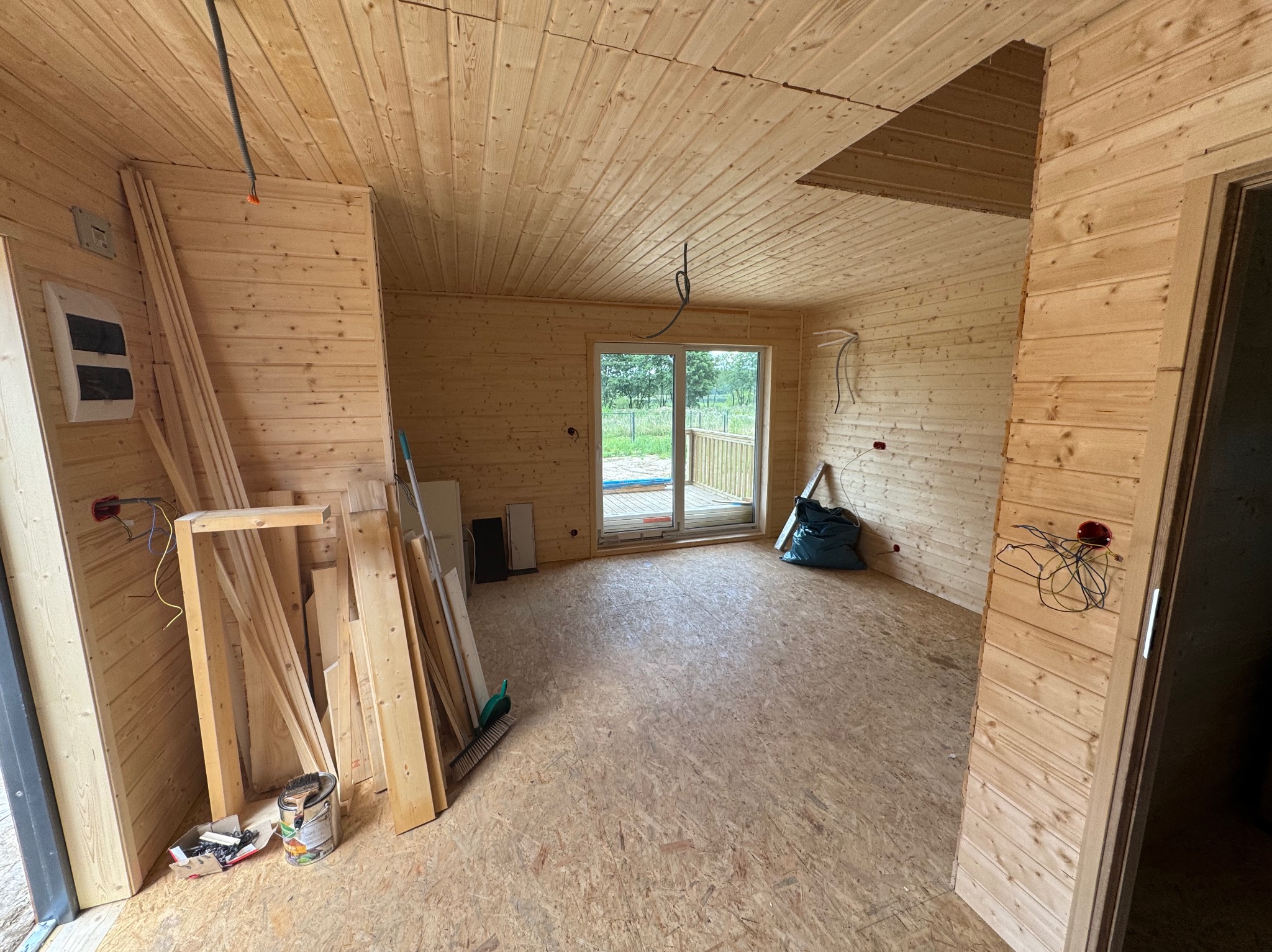 Wnętrze w trakcie budowy: drewniane ściany i sufit, widoczne instalacje elektryczne, okno balkonowe z widokiem na zewnątrz, narzędzia i materiały budowlane. Surowy stan wykończenia.