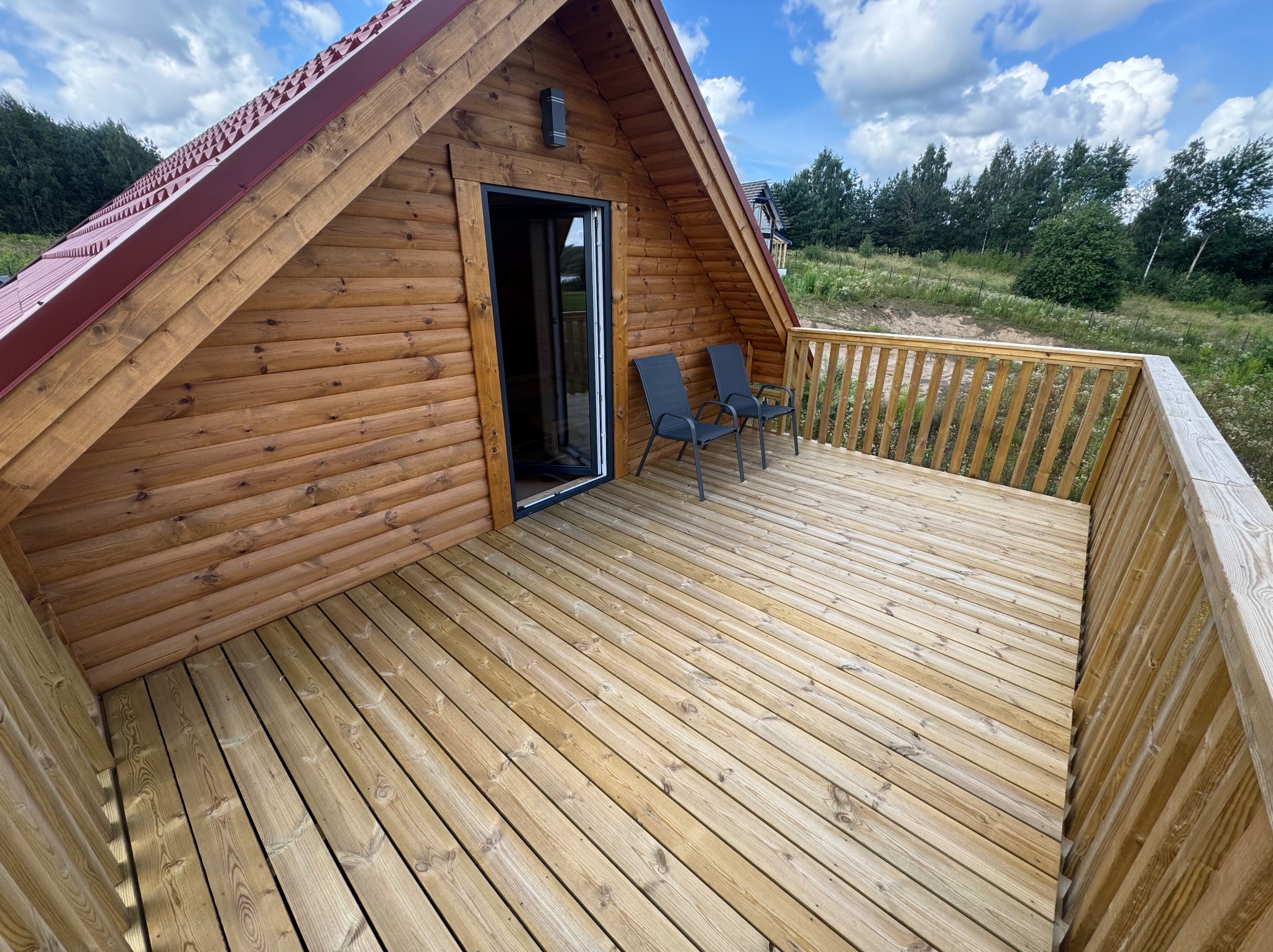 Drewniany domek z tarasem i dwoma krzesłami na tle zieleni i błękitnego nieba. Widok z góry, podkreślający strukturę drewna i przestrzeń tarasu.