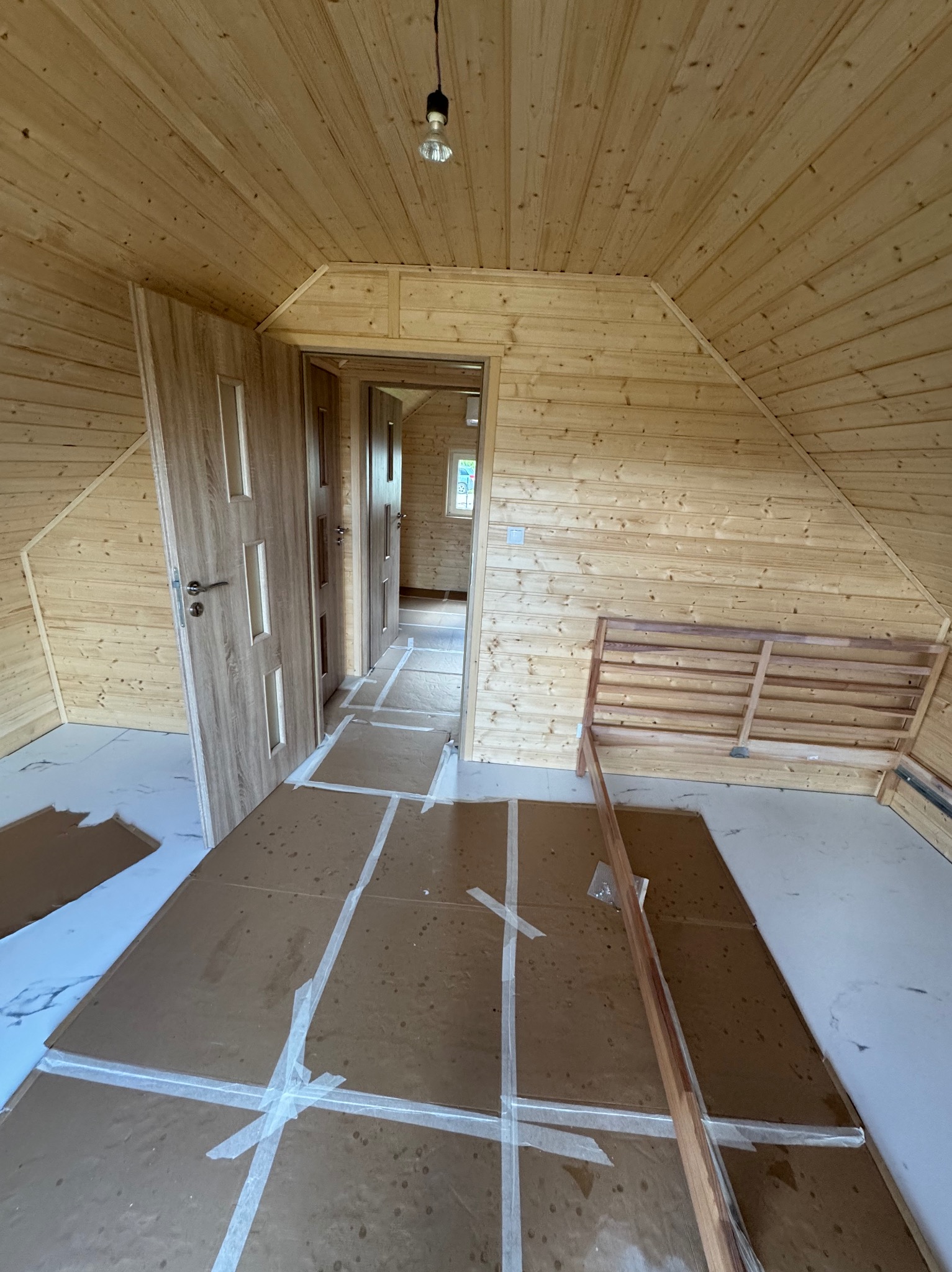 Wnętrze domu w budowie w Banino: drewniane ściany, podłoga zabezpieczona kartonem, widoczne drzwi i fragment konstrukcji. Surowy stan wykończenia.