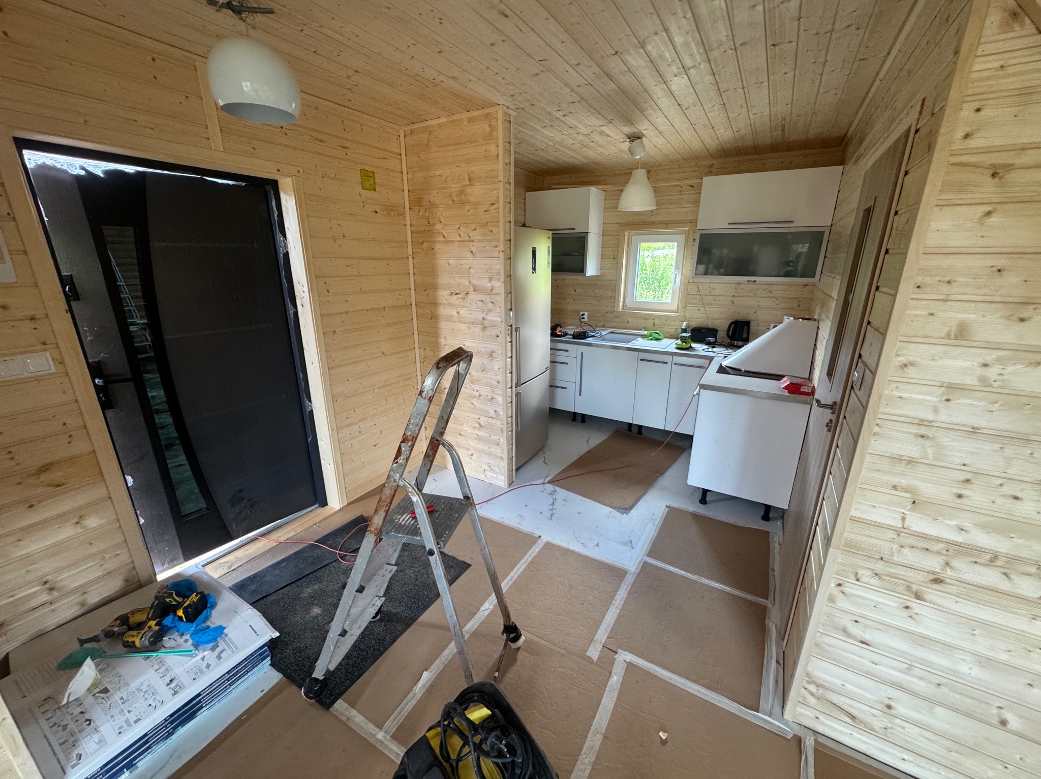 Wnętrze domu szkieletowego w trakcie wykańczania: drewniane ściany, zamontowana kuchnia, drabina i narzędzia na podłodze. Widok na okno i drzwi.