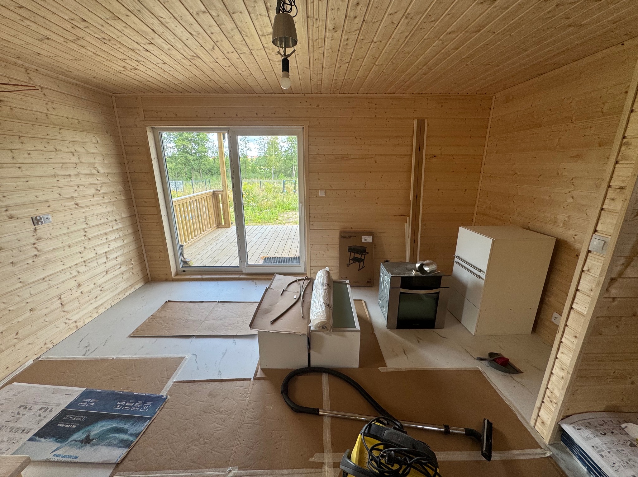 Wnętrze domu w trakcie wykańczania: drewniane ściany, okno balkonowe, częściowo położona podłoga, meble kuchenne, odkurzacz. Widoczne materiały budowlane.
