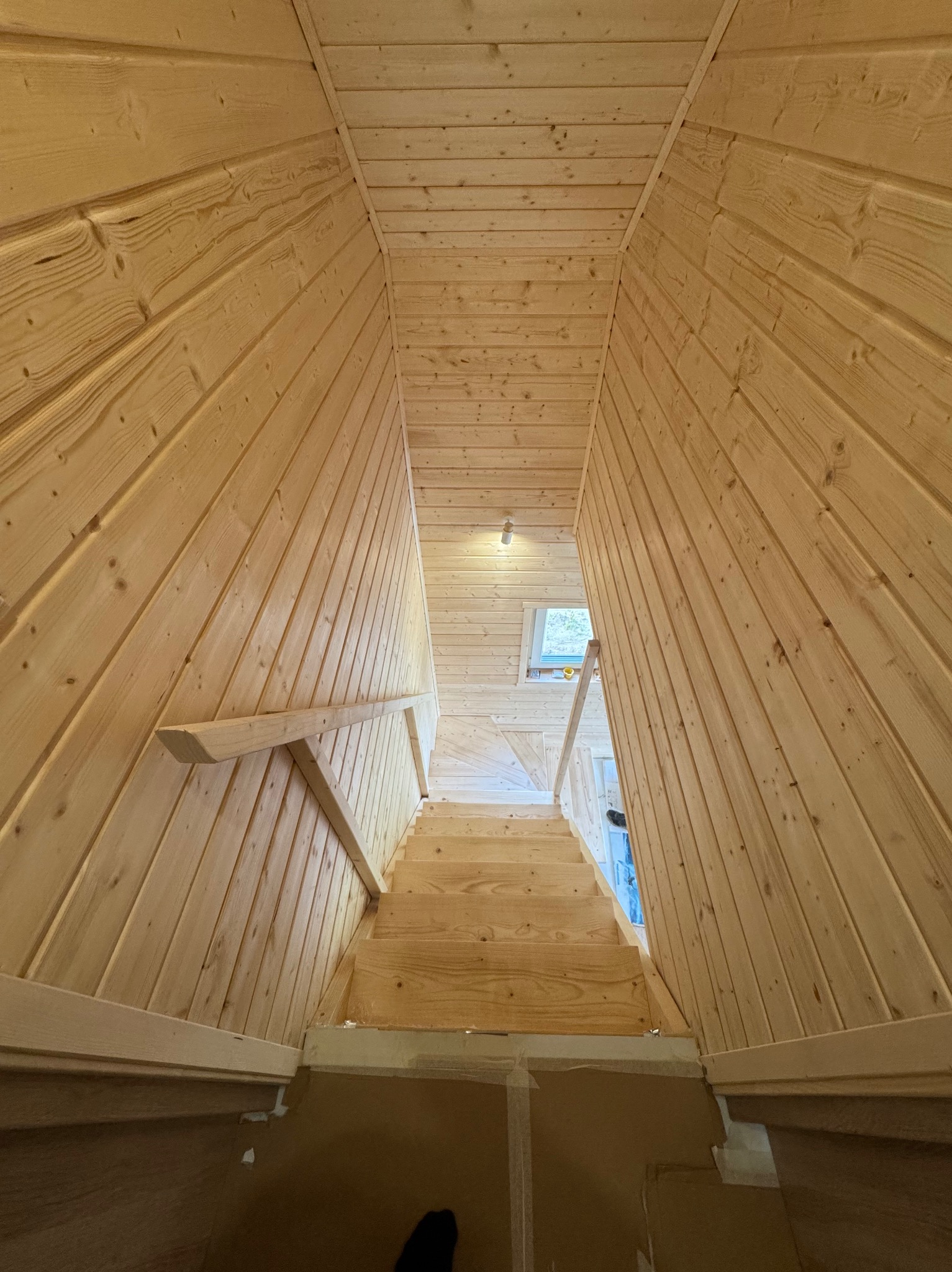 Wnętrze domu z drewnianymi schodami i boazerią. Ujęcie od dołu schodów, widoczne okno na końcu korytarza. Naturalne drewno, surowy stan wykończenia.