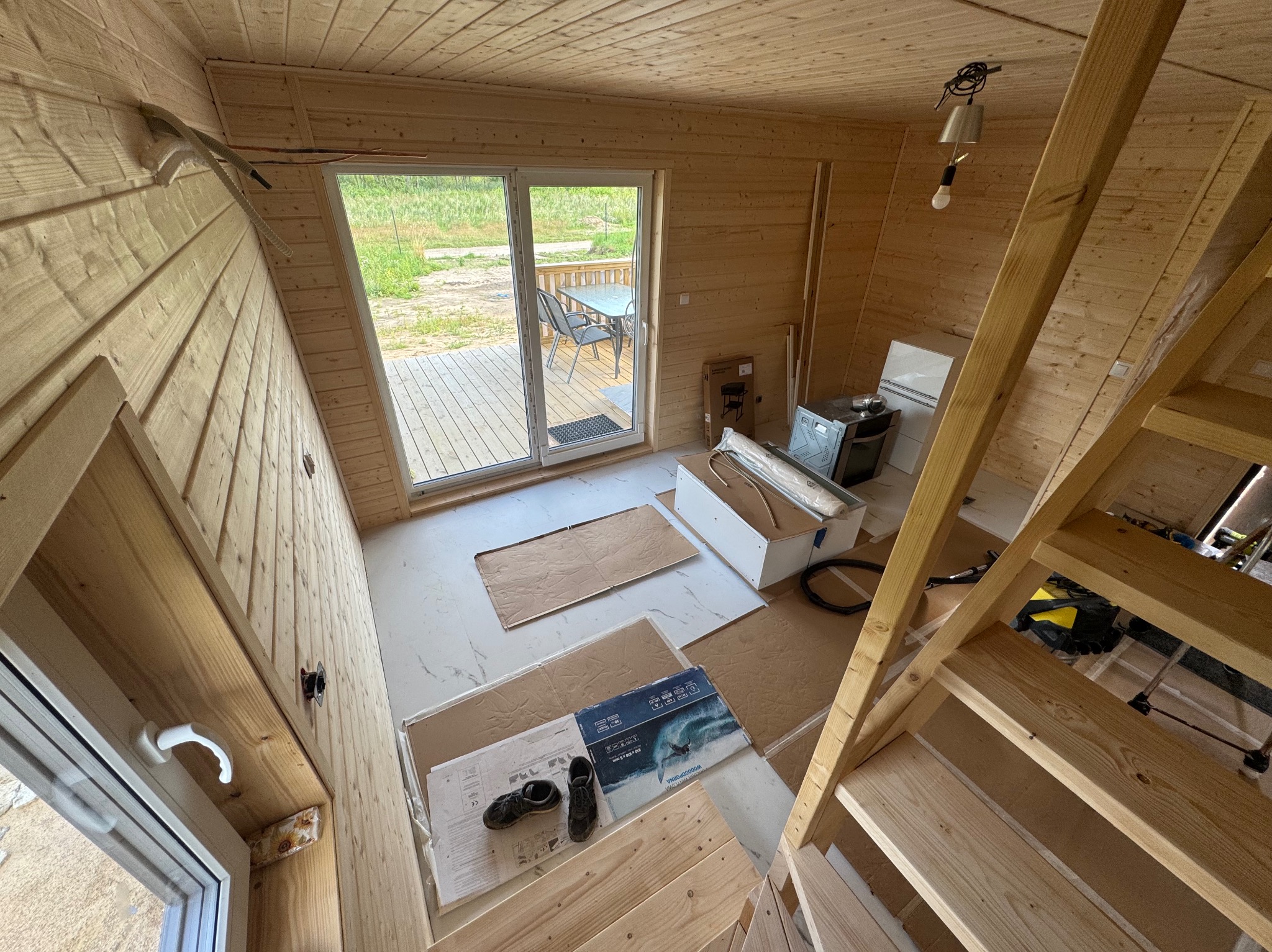 Wnętrze drewnianego domu w trakcie wykańczania, widoczne schody, okno z widokiem na zewnątrz, elementy wyposażenia i narzędzia. Charakterystyczna skośna ściana.