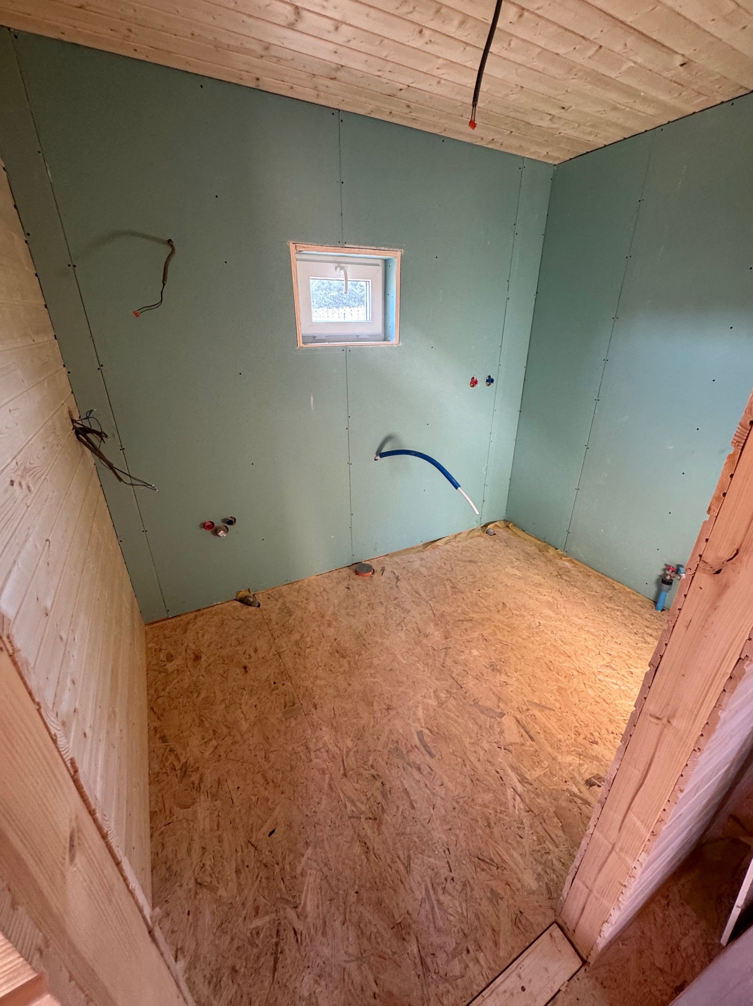 Wnętrze w trakcie budowy: płyty OSB na podłodze, ściany z płyt gipsowo-kartonowych, małe okno. Widoczne instalacje elektryczne i hydrauliczne.