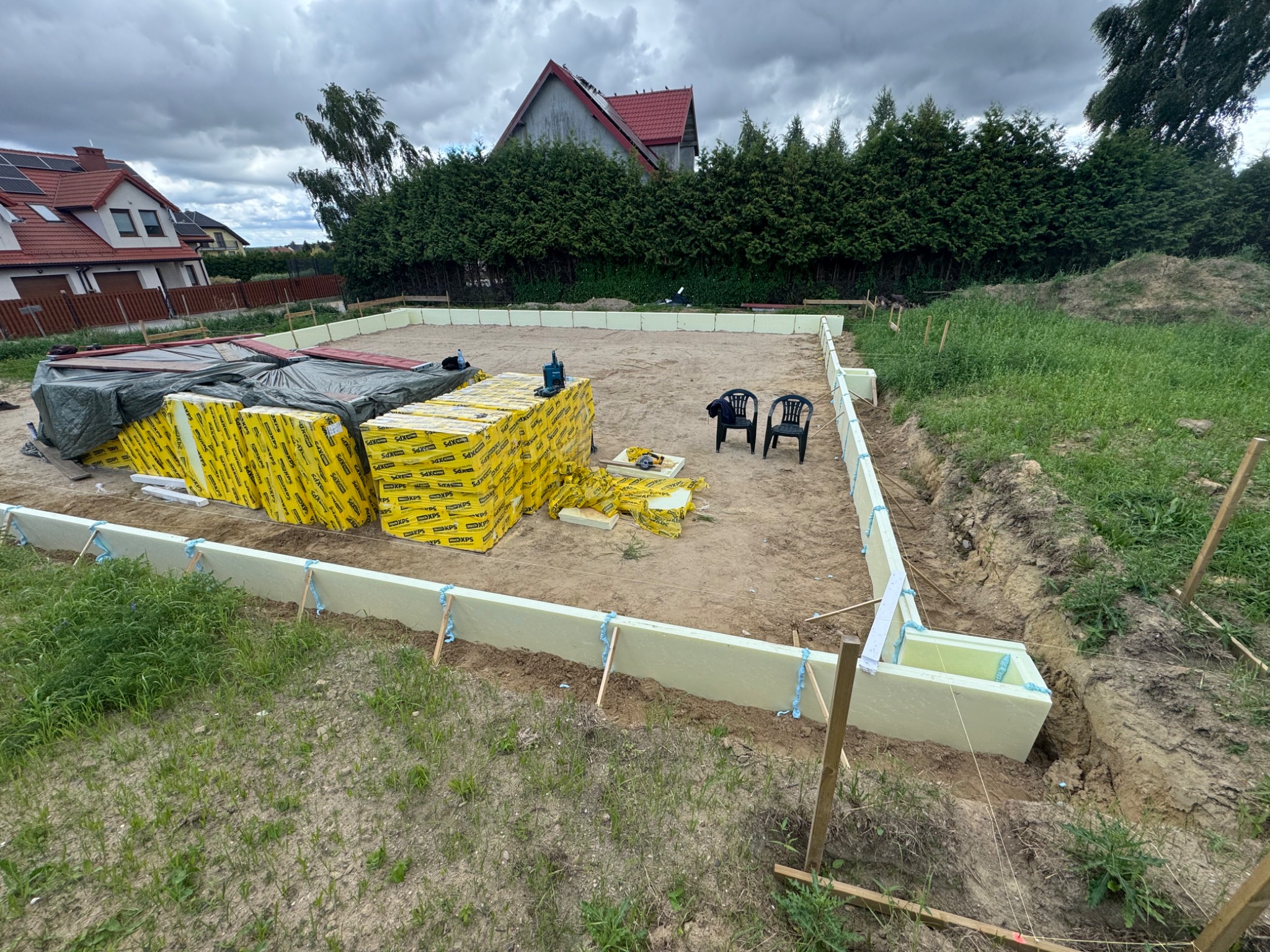 Przygotowanie fundamentów: izolacja termiczna ze styropianu XPS w wykopie pod budowę. Na działce leżą paczki materiałów budowlanych i narzędzia.