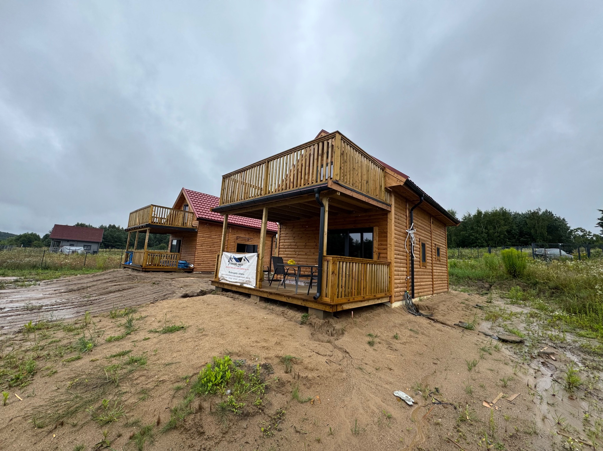 Dwa drewniane domy z balkonami, jeden z czerwoną dachówką, na piaszczystym terenie, pochmurne niebo. Widoczny baner reklamowy na jednym z domów.