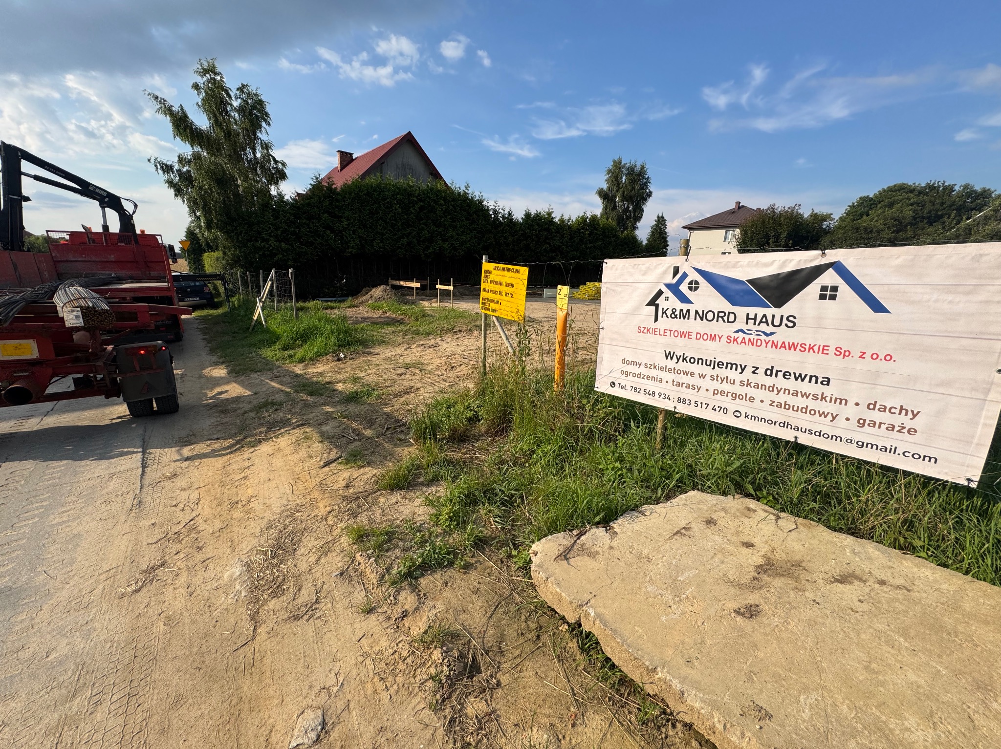 Teren budowy domu szkieletowego z tablicą reklamową K&M Nord Haus, widoczny transport materiałów budowlanych. W tle zieleń i domy.