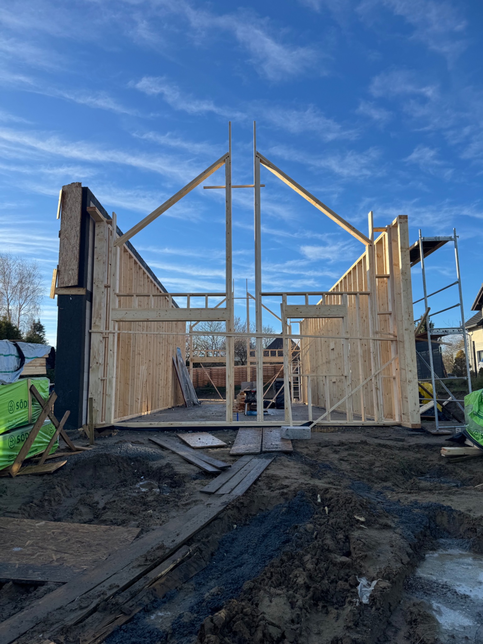 Wznoszenie konstrukcji szkieletowej domu jednorodzinnego w słoneczny dzień. Widoczne drewniane elementy konstrukcyjne i rusztowanie. Na pierwszym planie ziemia.