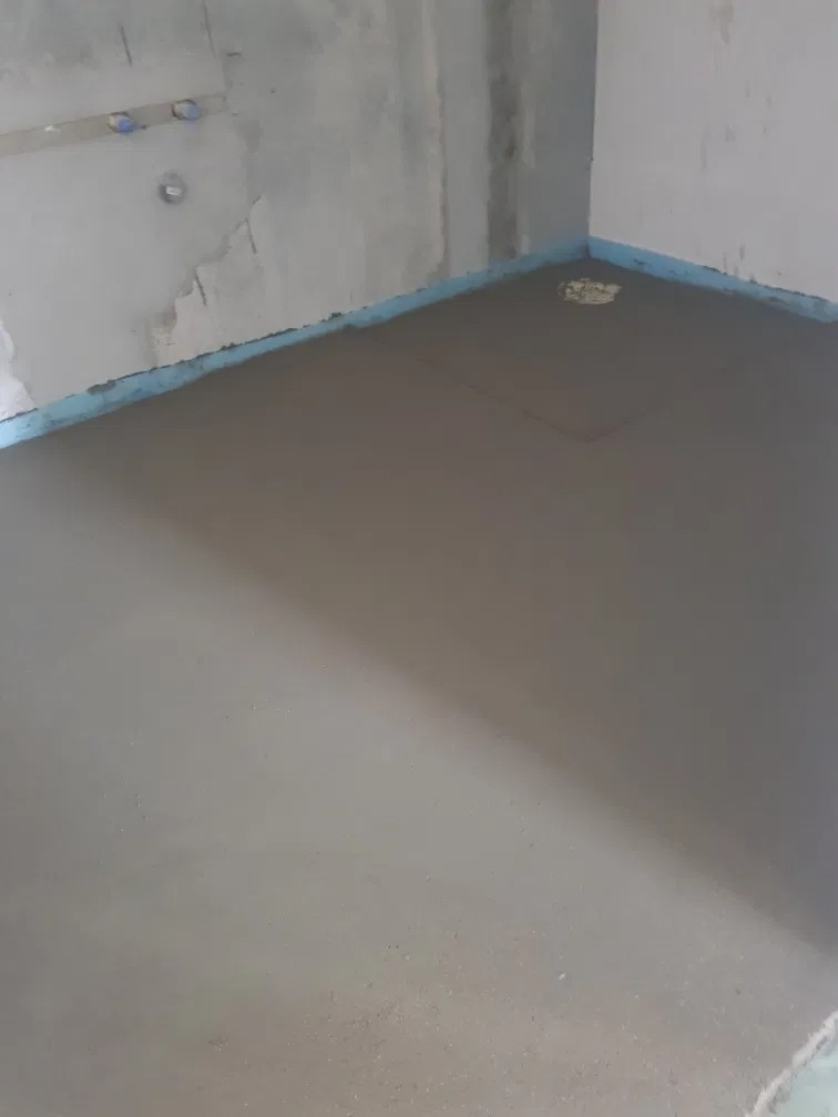 Świeżo wylana, gładka posadzka w szarym odcieniu, zabezpieczona niebieską taśmą przy ścianach, z widocznymi śladami po narzędziach do rozprowadzania masy.