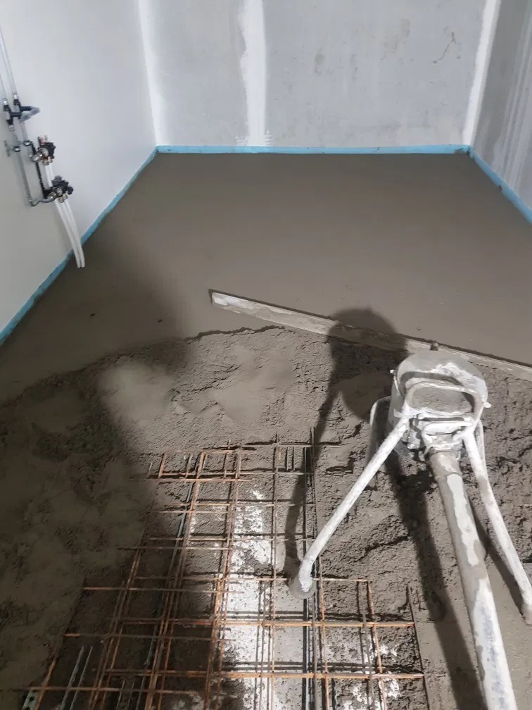 Wnętrze w trakcie wylewania posadzki: widoczna siatka zbrojeniowa, wylewka w trakcie rozprowadzania maszyną, częściowo wyrównana powierzchnia, instalacja wodna na ścianie.
