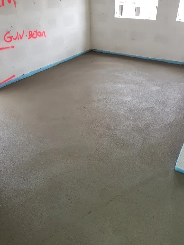 Świeżo wylana, gładka posadzka w pomieszczeniu w stanie surowym, zabezpieczona taśmą przy ścianach, z widocznym napisem 'Gulv Beton' na ścianie.