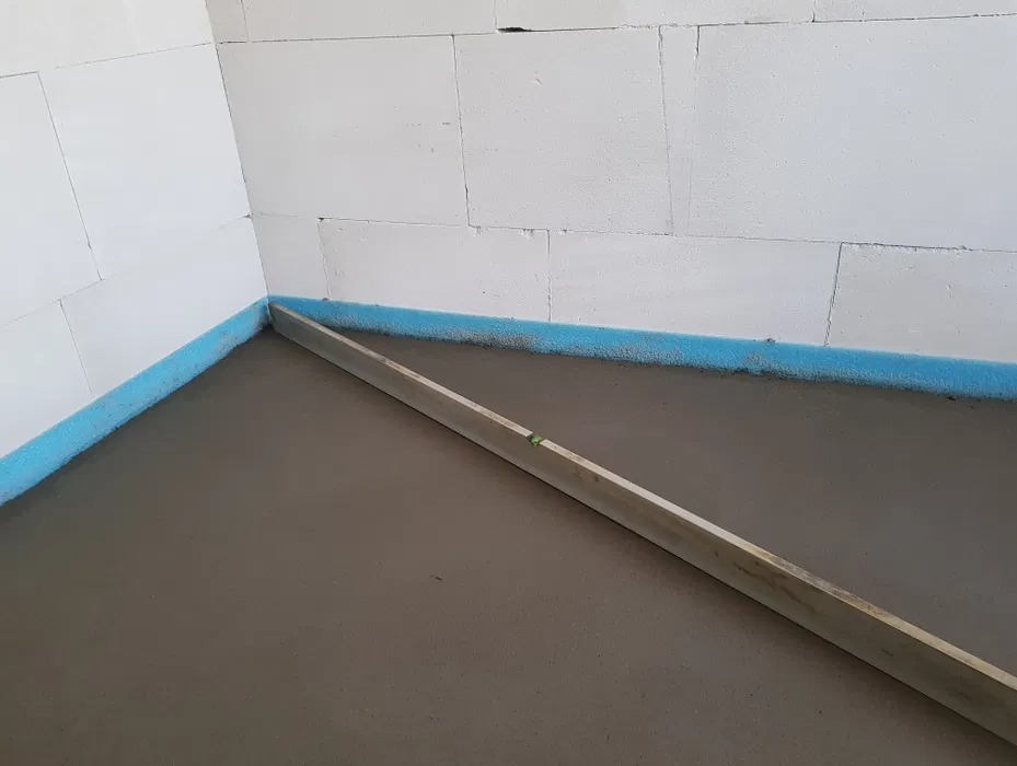Świeżo wylana, szara posadzka w pomieszczeniu z białymi ścianami z bloczków, zabezpieczona niebieską pianką przy ścianach i wyrównywana długą, drewnianą łatą.