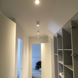 SIGMA ENGINEERING SPÓŁKA Z OGRANICZONĄ ODPOWIEDZIALNOŚCIĄ - Korytarz w trakcie remontu z zamontowanymi punktowymi lampami sufitowymi, widoczne otwarte szafy wnękowe i pracownik w oddali.