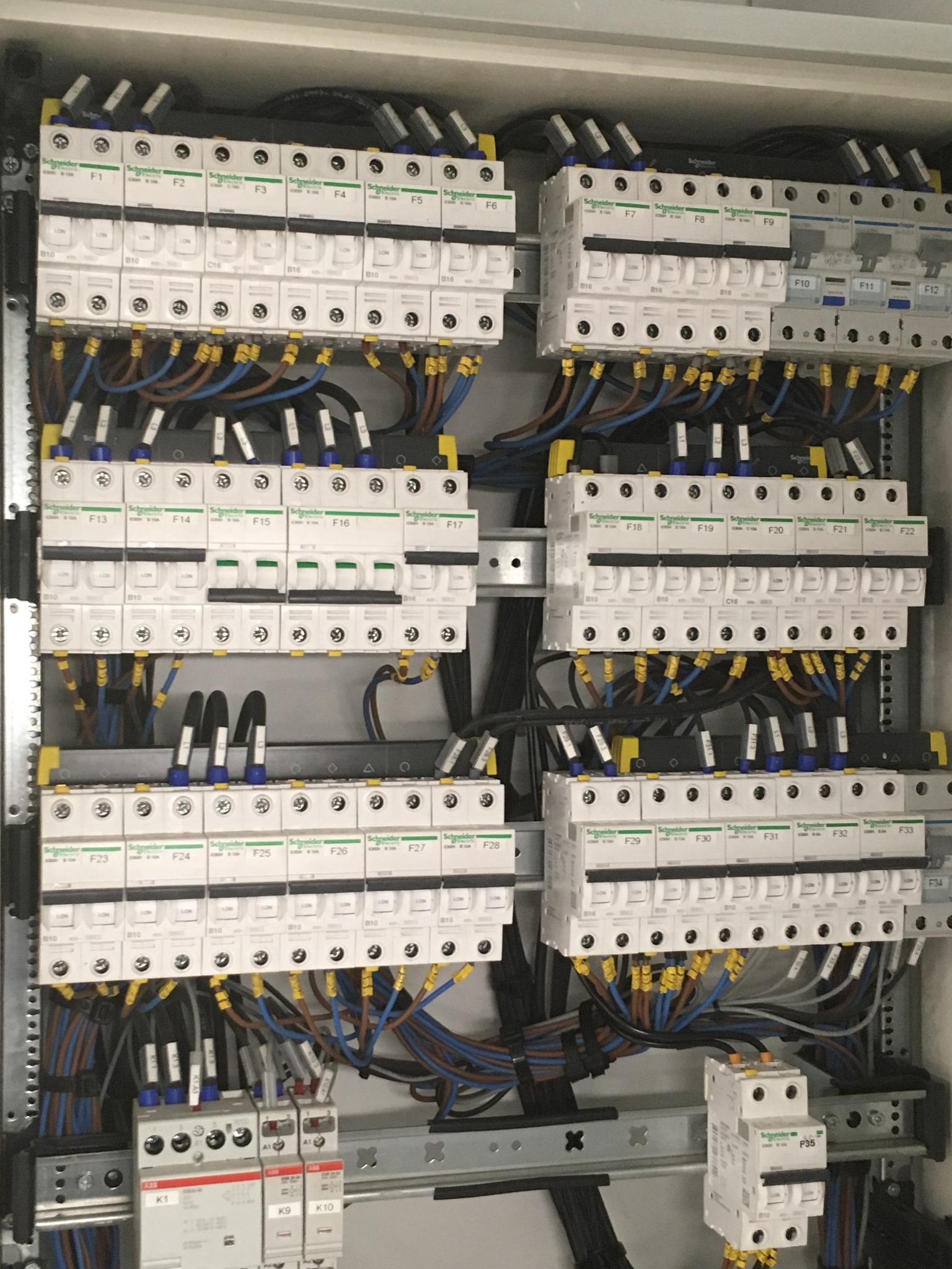 Rozdzielnica elektryczna z zainstalowanymi bezpiecznikami i okablowaniem, widoczne elementy marki Schneider Electric, ułożone w rzędach i kolumnach wewnątrz metalowej obudowy.