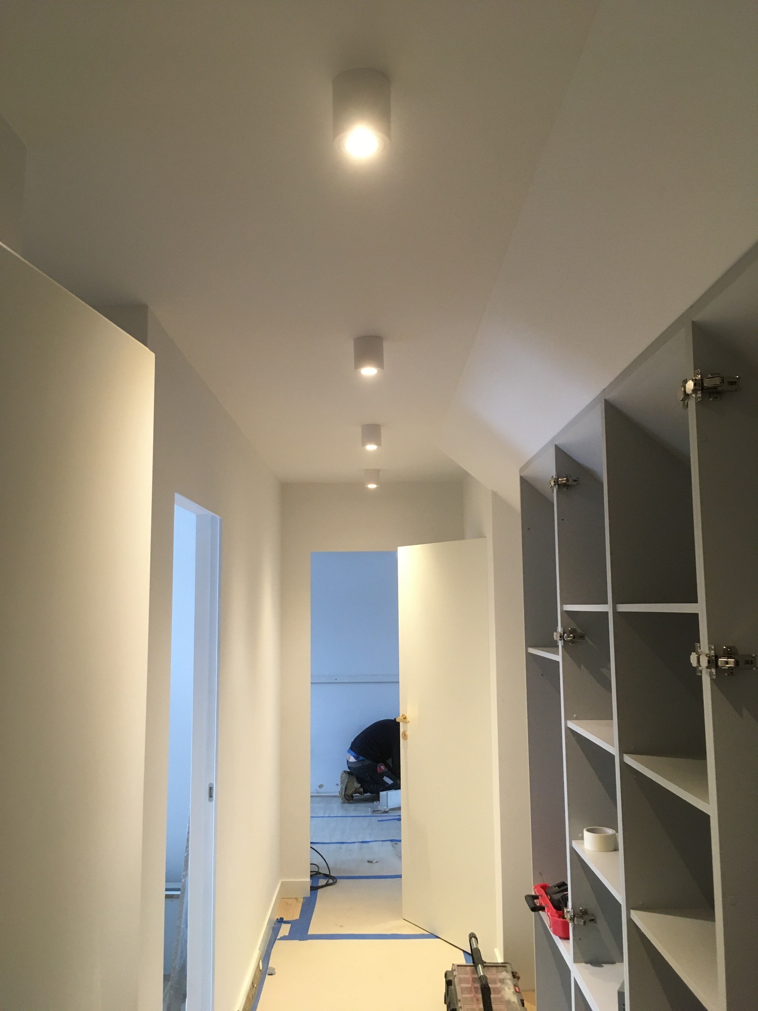 Korytarz w trakcie remontu z zamontowanymi punktowymi lampami sufitowymi, widoczne otwarte szafy wnękowe i pracownik w oddali.