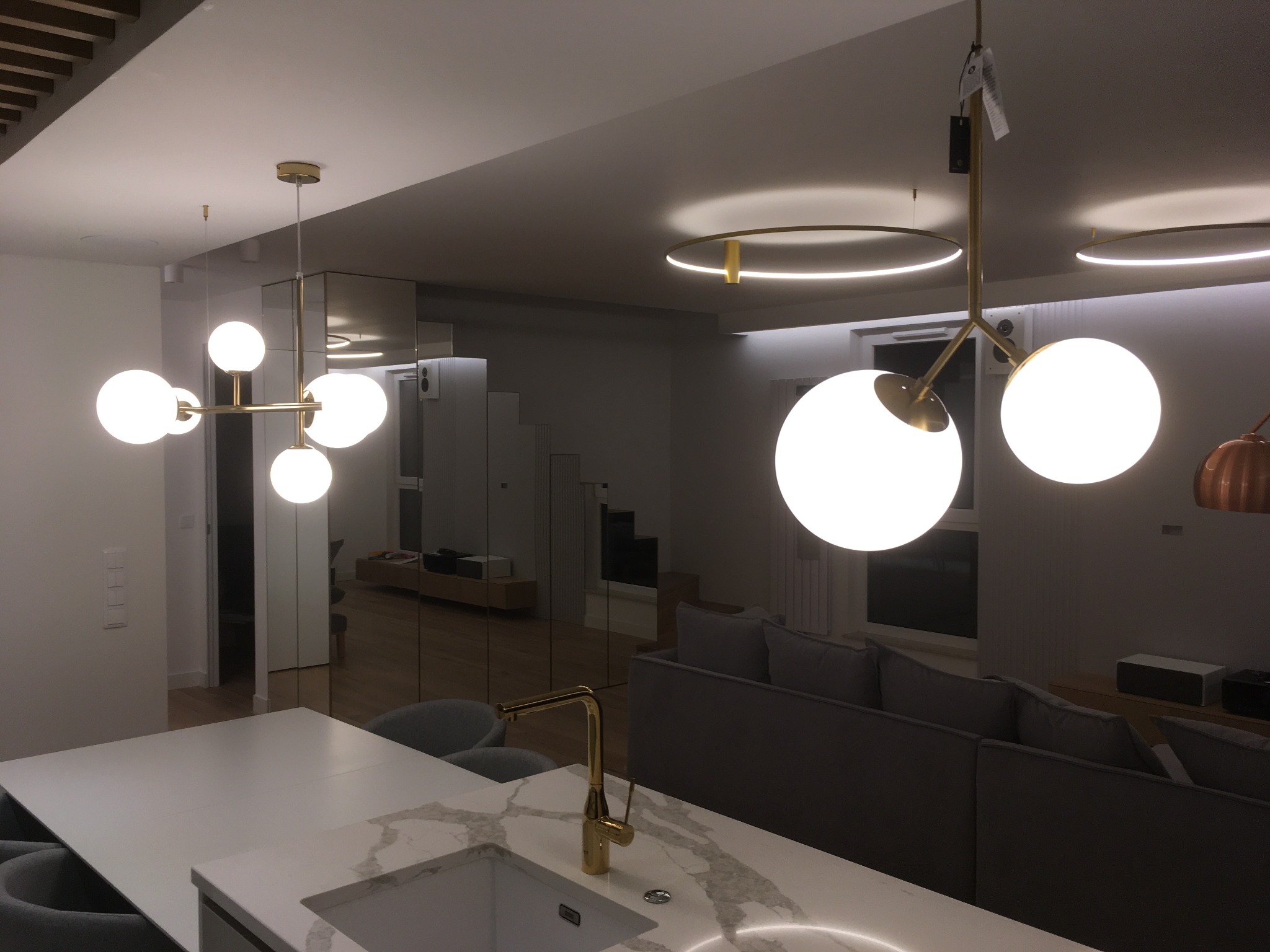 Nowoczesne oświetlenie wnętrza: kuliste lampy wiszące i okrągłe plafony LED w salonie z wyspą kuchenną i złotą armaturą.