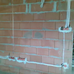 Instalacja elektryczna w trakcie montażu w nowym budynku z cegły, widoczne przewody i puszki elektryczne zamocowane w ścianie. Częściowo wykonane bruzdy pod kable.
