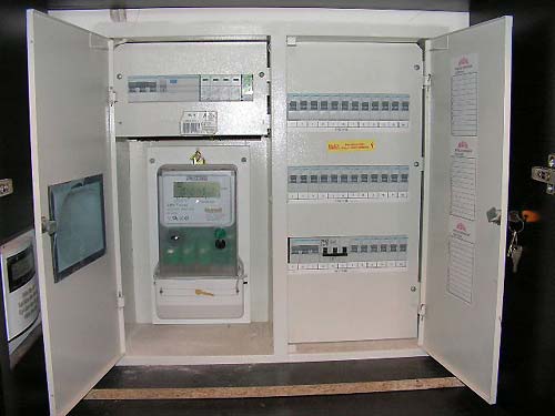 Otwarta szafka rozdzielcza z licznikiem energii elektrycznej i bezpiecznikami, widoczne klucze w zamku drzwi.