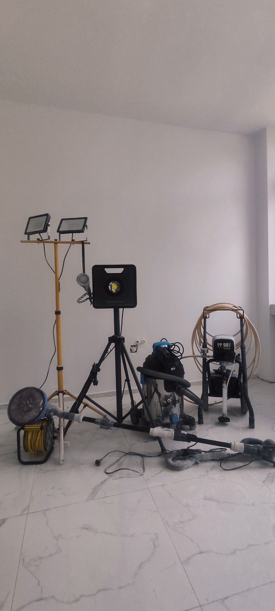 Sprzęt do szlifowania i malowania ścian: szlifierka z odkurzaczem, agregat malarski PP 90, lampy budowlane ScanGrip na statywach w pustym pomieszczeniu z białymi ścianami i marmurową podłogą.