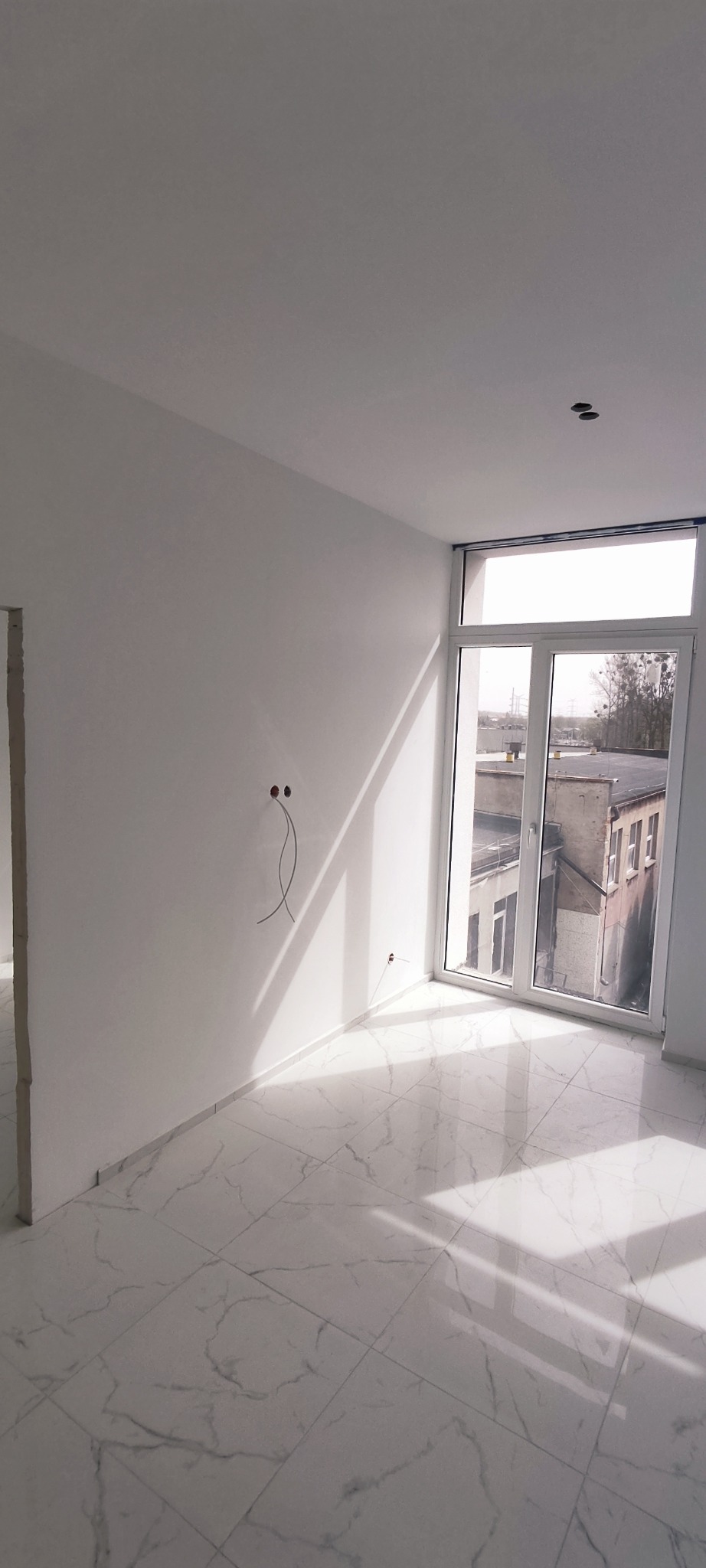 Pomalowane na biało wnętrze z marmurową podłogą, widoczne wystające kable ze ściany i jasne światło wpadające przez okno.