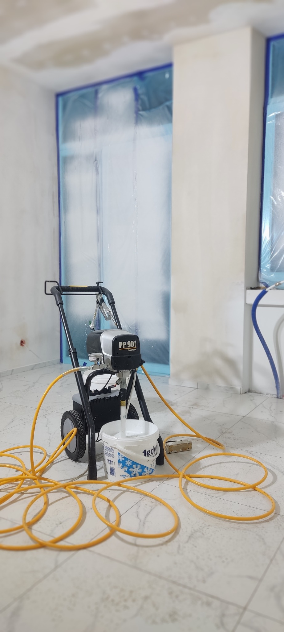 Agregat malarski PP 90 Extra z żółtym wężem i wiadrem farby na marmurowej podłodze, okna i framugi zabezpieczone folią malarską, przygotowanie do malowania.