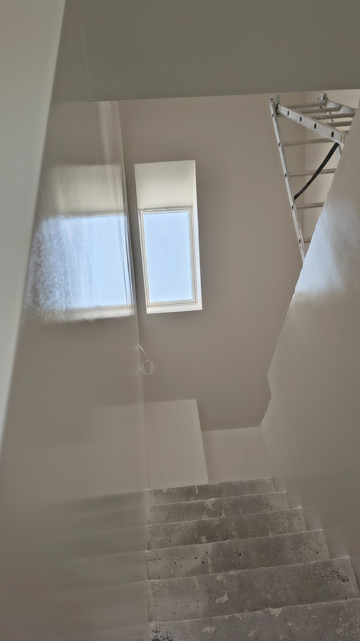 Świeżo pomalowane na biało ściany klatki schodowej z oknem dachowym i aluminiową drabiną opartą o ścianę, widoczne surowe betonowe schody.