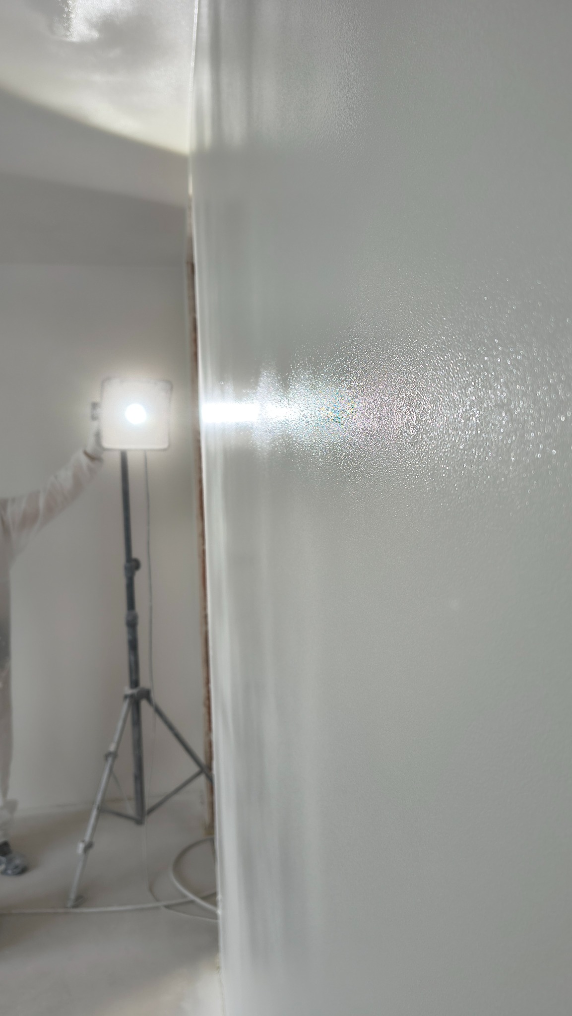 Świeżo pomalowana, biała ściana z widocznymi refleksami światła lampy na statywie i fragmentem osoby w kombinezonie ochronnym.
