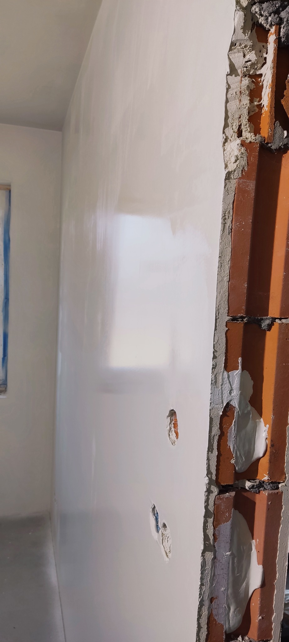 Świeżo położona, lśniąca gładź na ścianie z widocznymi śladami po łataniu ubytków, przylegająca do surowego muru z cegły z pozostałościami zaprawy.