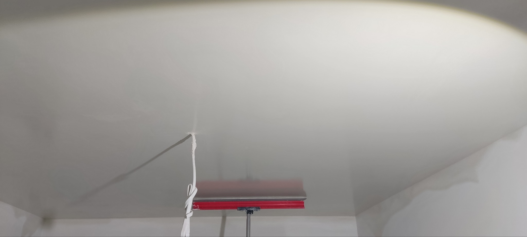 Gładka, świeżo wykończona powierzchnia sufitu z widocznym narzędziem do rozprowadzania gładzi na teleskopowym uchwycie oraz sznurkiem zwisającym z sufitu.