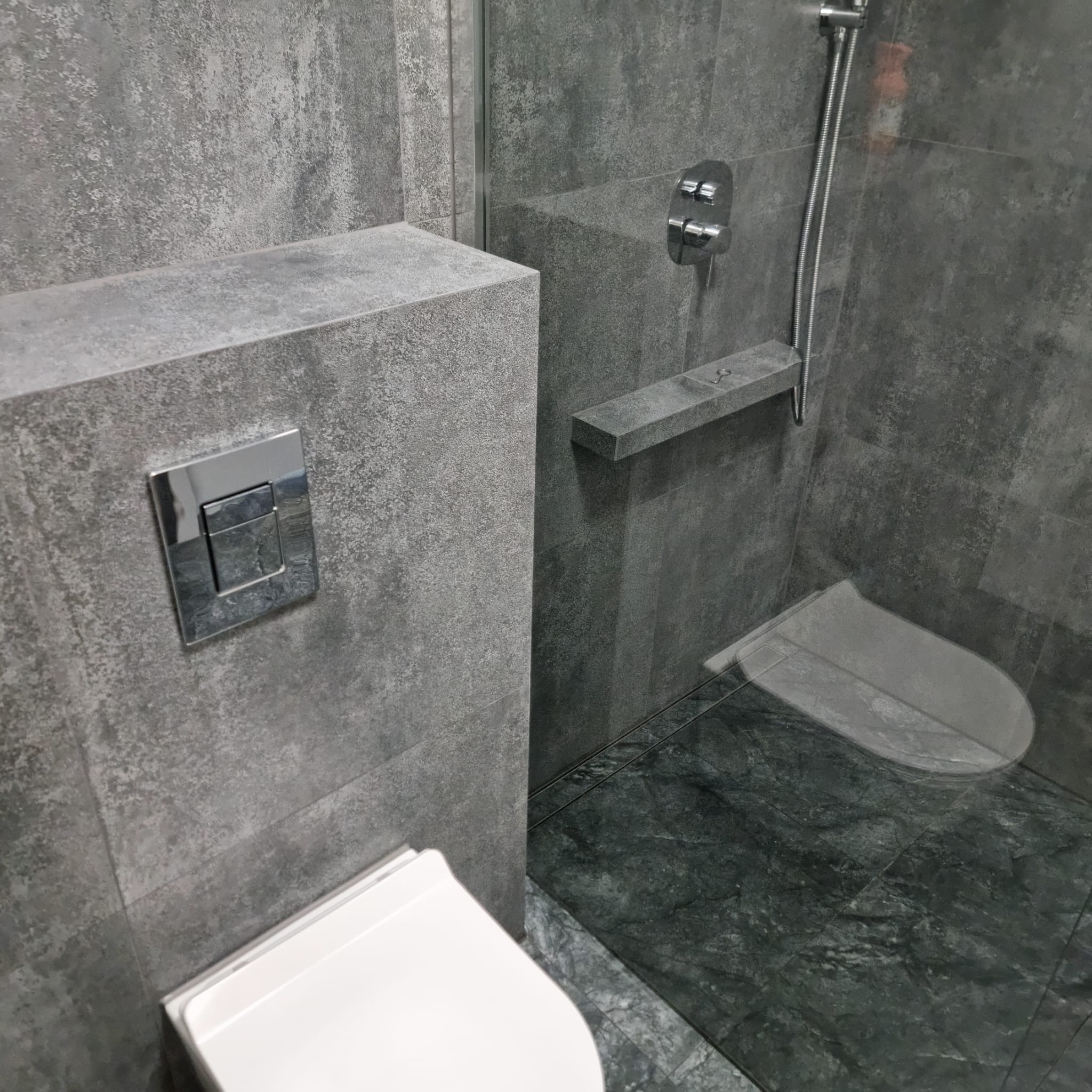 Nowoczesna łazienka w odcieniach szarości z podtynkową spłuczką i kabiną prysznicową typu walk-in z chromowaną armaturą oraz zieloną posadzką.