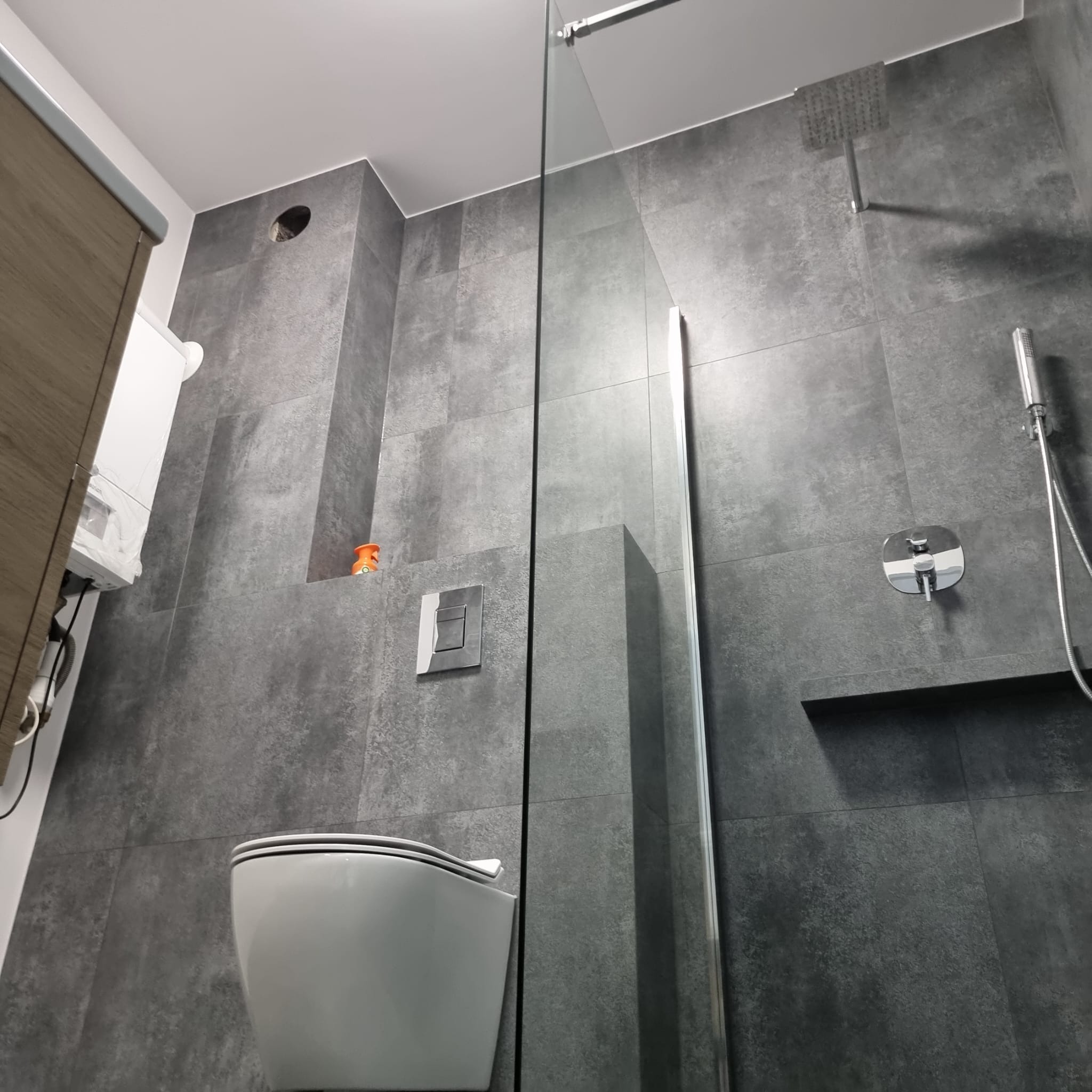 Nowoczesna łazienka z szarymi płytkami imitującymi beton, kabiną prysznicową typu walk-in, podtynkową toaletą i deszczownicą.