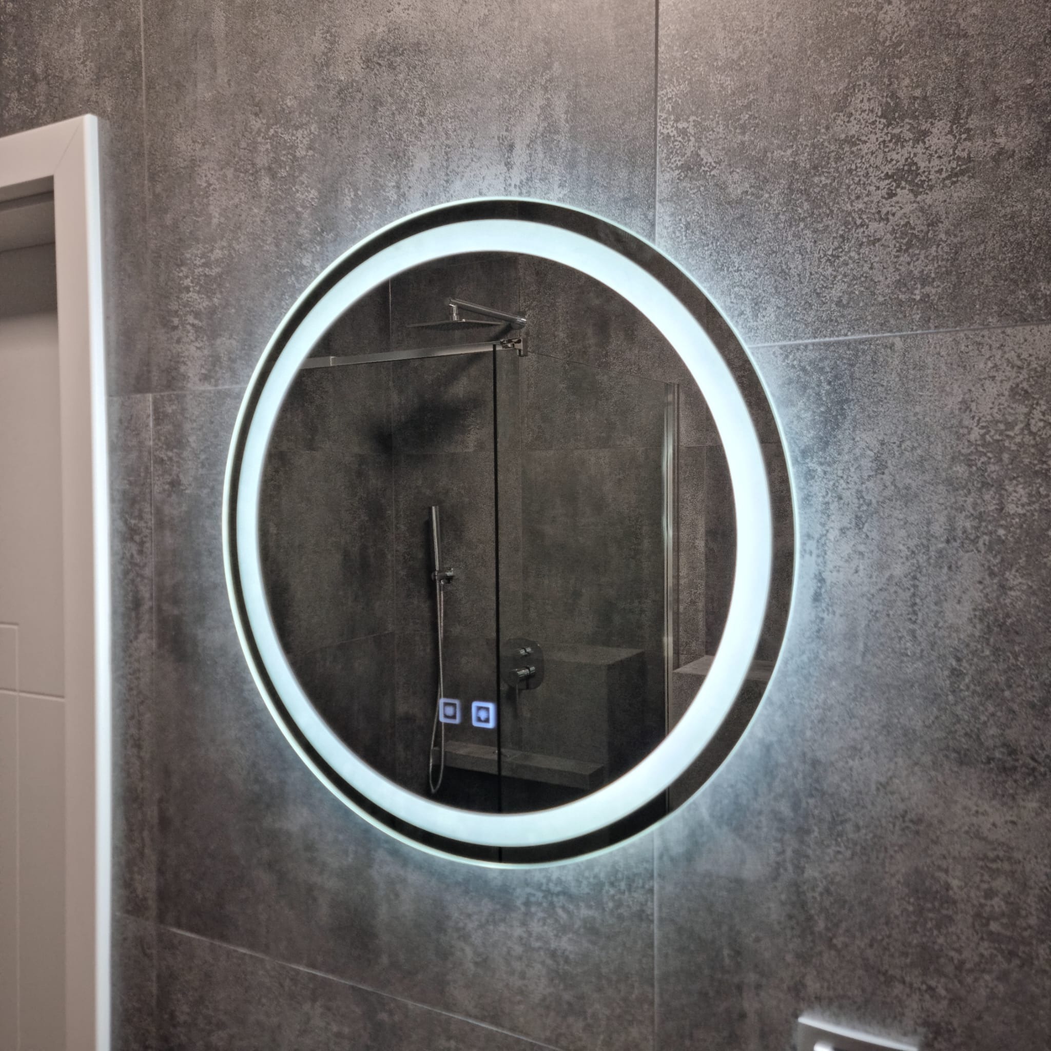 Okrągłe lustro z podświetleniem LED na szarej, teksturowanej ścianie w łazience, odbijające kabinę prysznicową z deszczownicą i panelem sterowania.