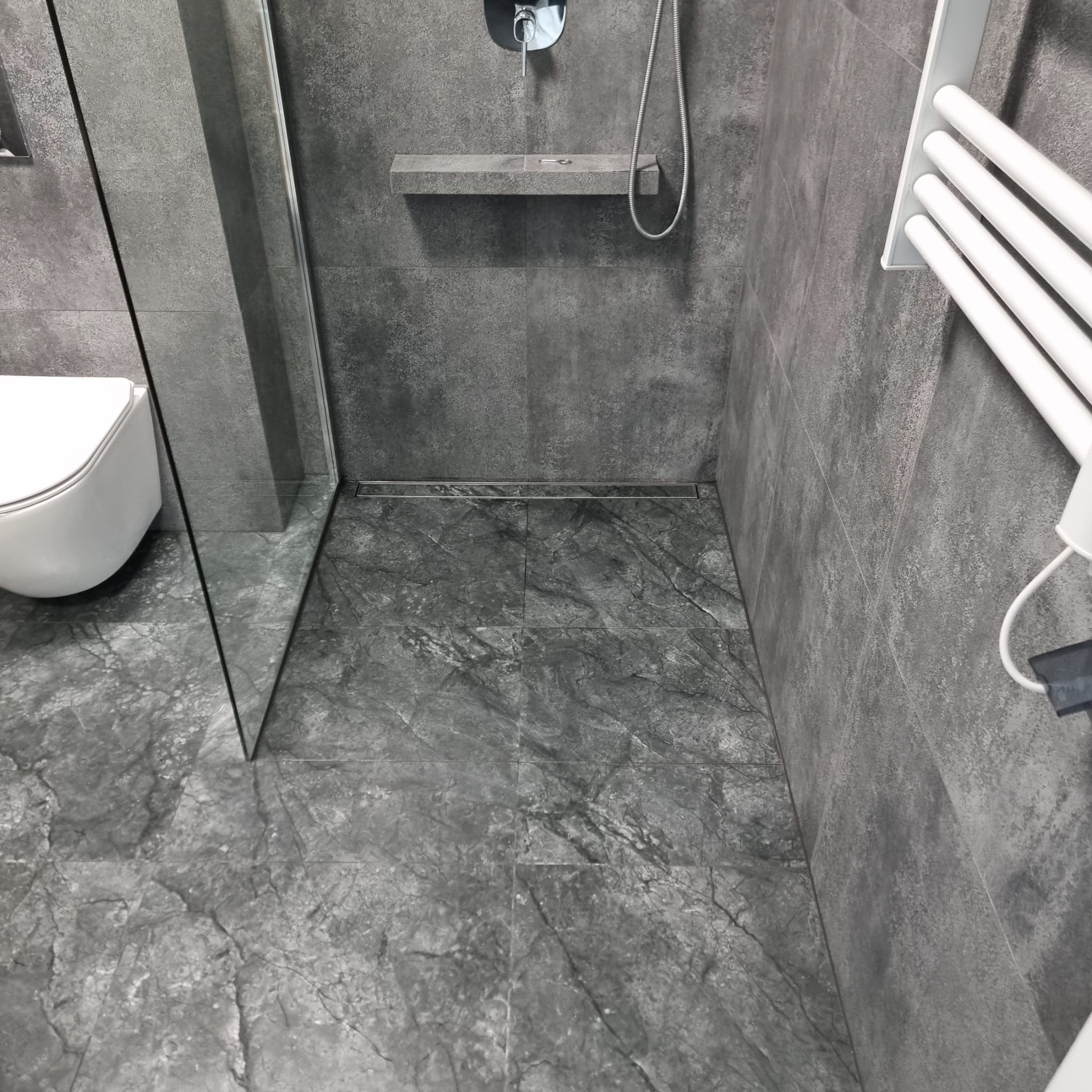 Szara łazienka z kabiną prysznicową typu walk-in, odpływem liniowym w podłodze, wiszącą toaletą i grzejnikiem drabinkowym.