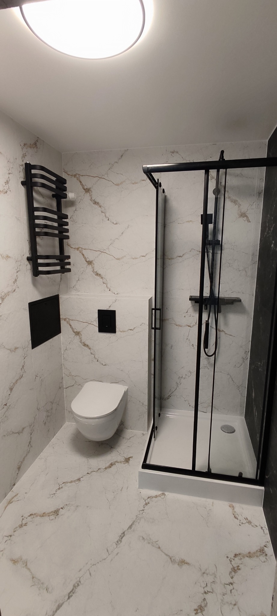 Nowoczesna łazienka z kabiną prysznicową typu walk-in z czarnymi profilami, wiszącą toaletą i czarnym grzejnikiem drabinkowym na ścianie z marmurowym wzorem.