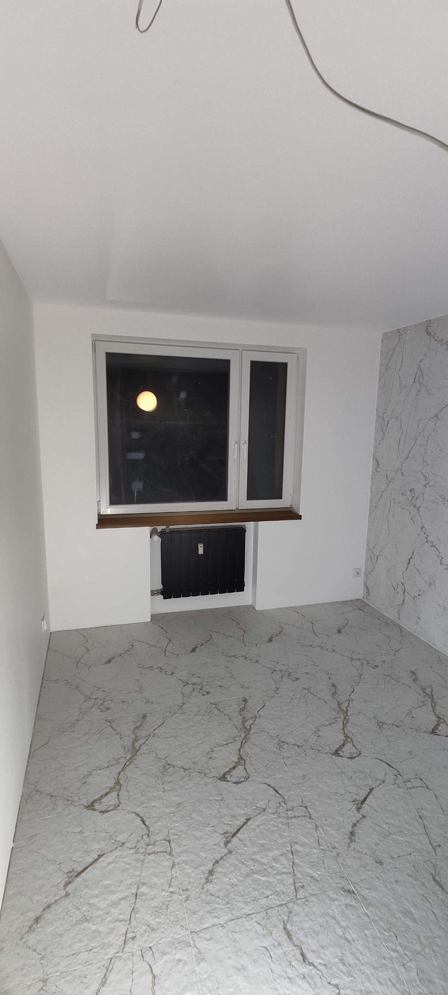 Pusty pokój po remoncie z marmurową podłogą, świeżo pomalowanymi ścianami, grzejnikiem pod oknem i tapetą w marmurkowy wzór na jednej ze ścian.