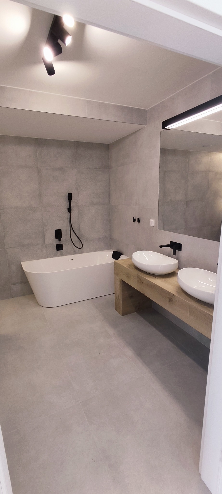 Nowoczesna łazienka z szarymi płytkami, wolnostojącą białą wanną, drewnianą szafką pod umywalki nablatowe i czarną armaturą.