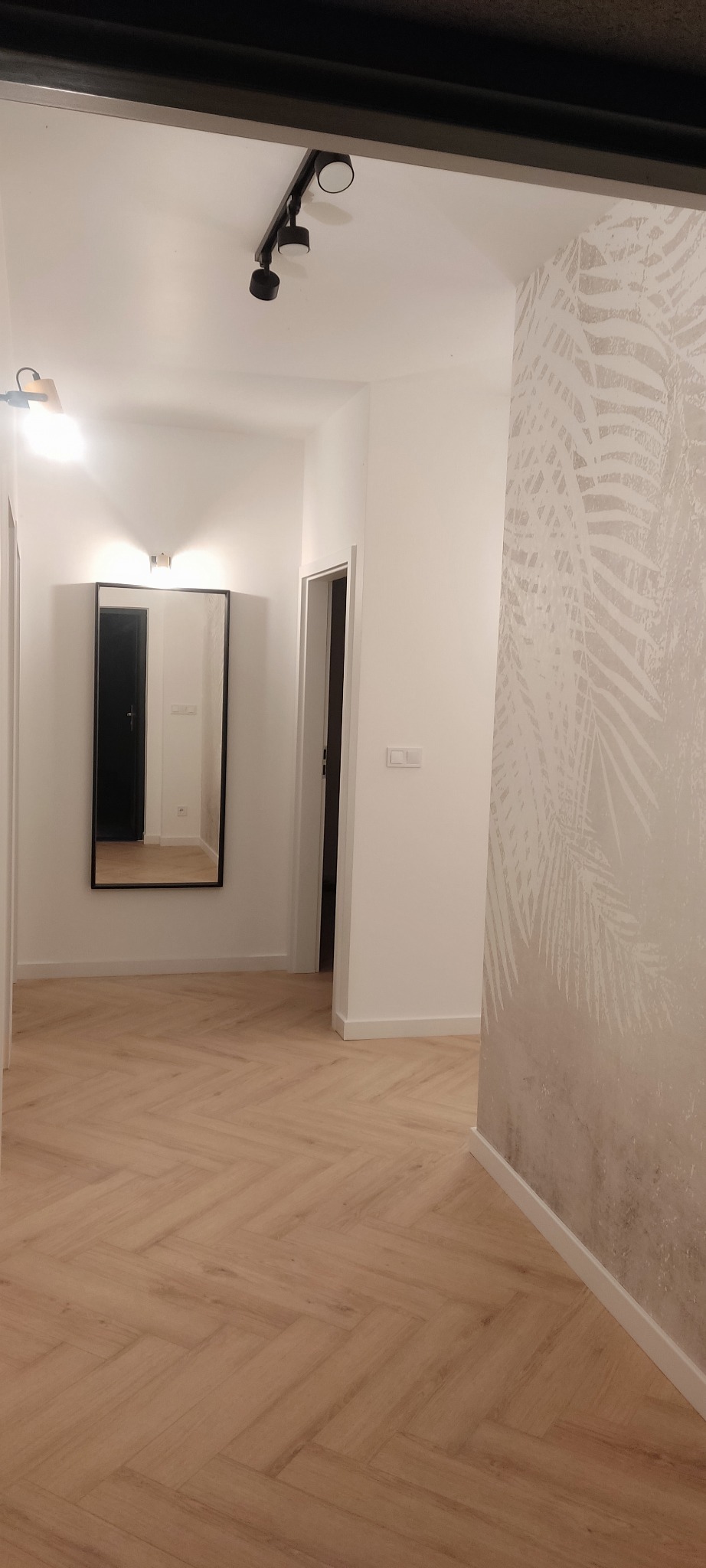 Nowoczesny przedpokój z parkietem ułożonym w jodełkę, lustrem w czarnej ramie i tapetą w motywy roślinne.