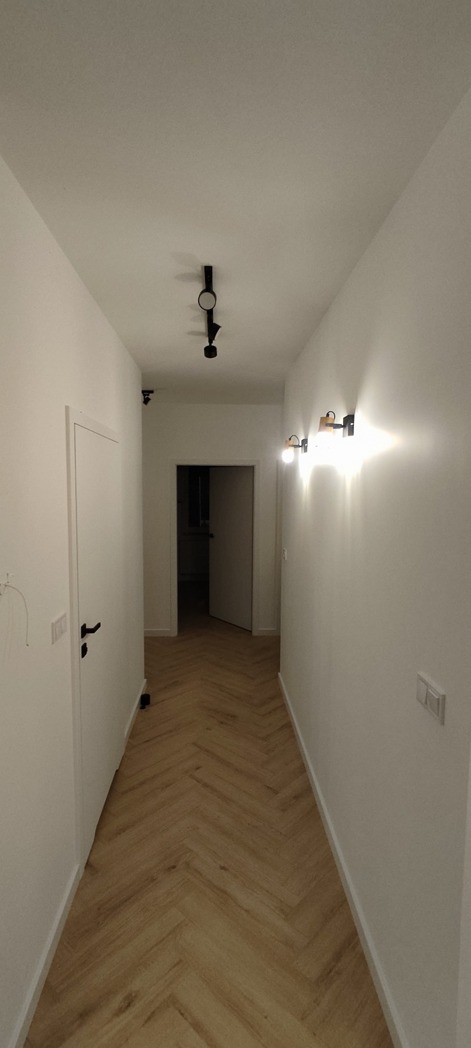 Korytarz po remoncie z parkietem ułożonym w jodełkę, białymi ścianami z zamontowanymi czarnymi lampami i białymi drzwiami z czarną klamką.