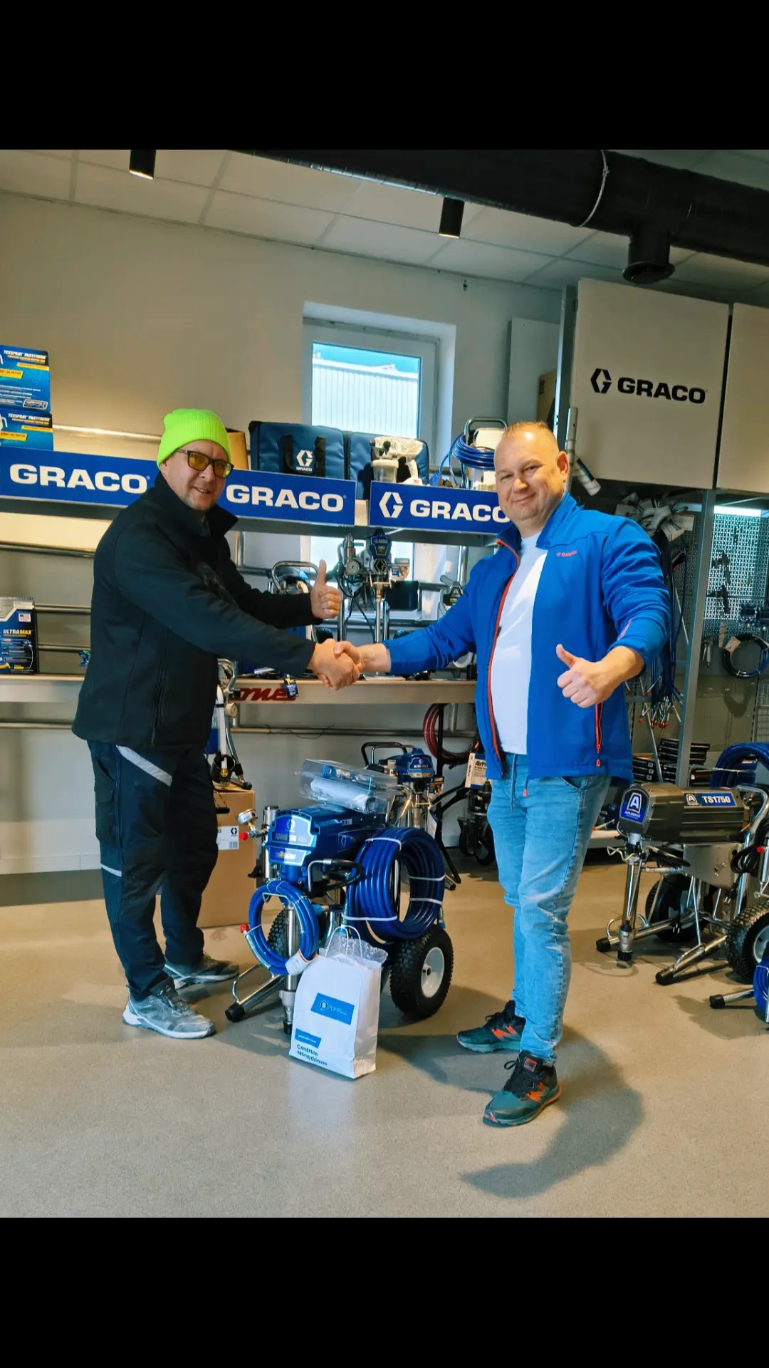 Dwóch mężczyzn w sklepie z urządzeniami malarskimi Graco, jeden w neonowej czapce, podaje sobie ręce nad nowym agregatem malarskim na kółkach.