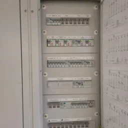 Otwarta skrzynka rozdzielcza z bezpiecznikami i schematami elektrycznymi, widoczne oznaczenia i przełączniki, szara obudowa.