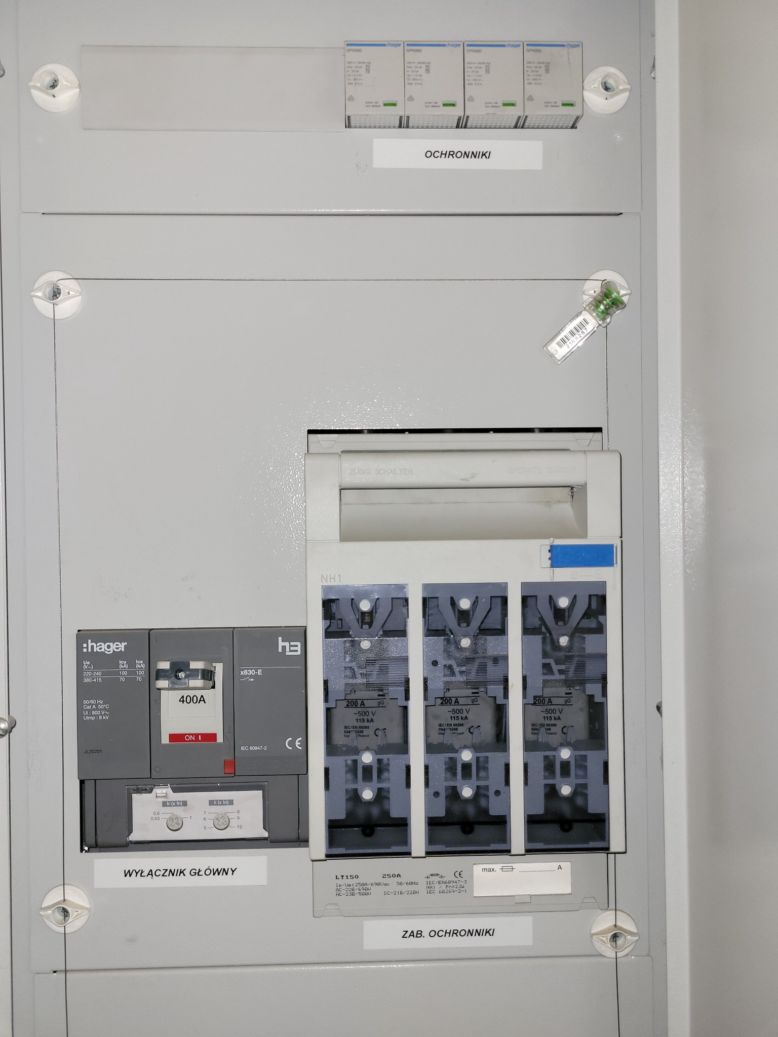 Szara skrzynka elektryczna z widocznymi bezpiecznikami, wyłącznikiem głównym Hager 400A i zabezpieczeniami topikowymi 200A, z naklejkami informacyjnymi 'Ochronniki' i 'Wyłącznik Główny'.