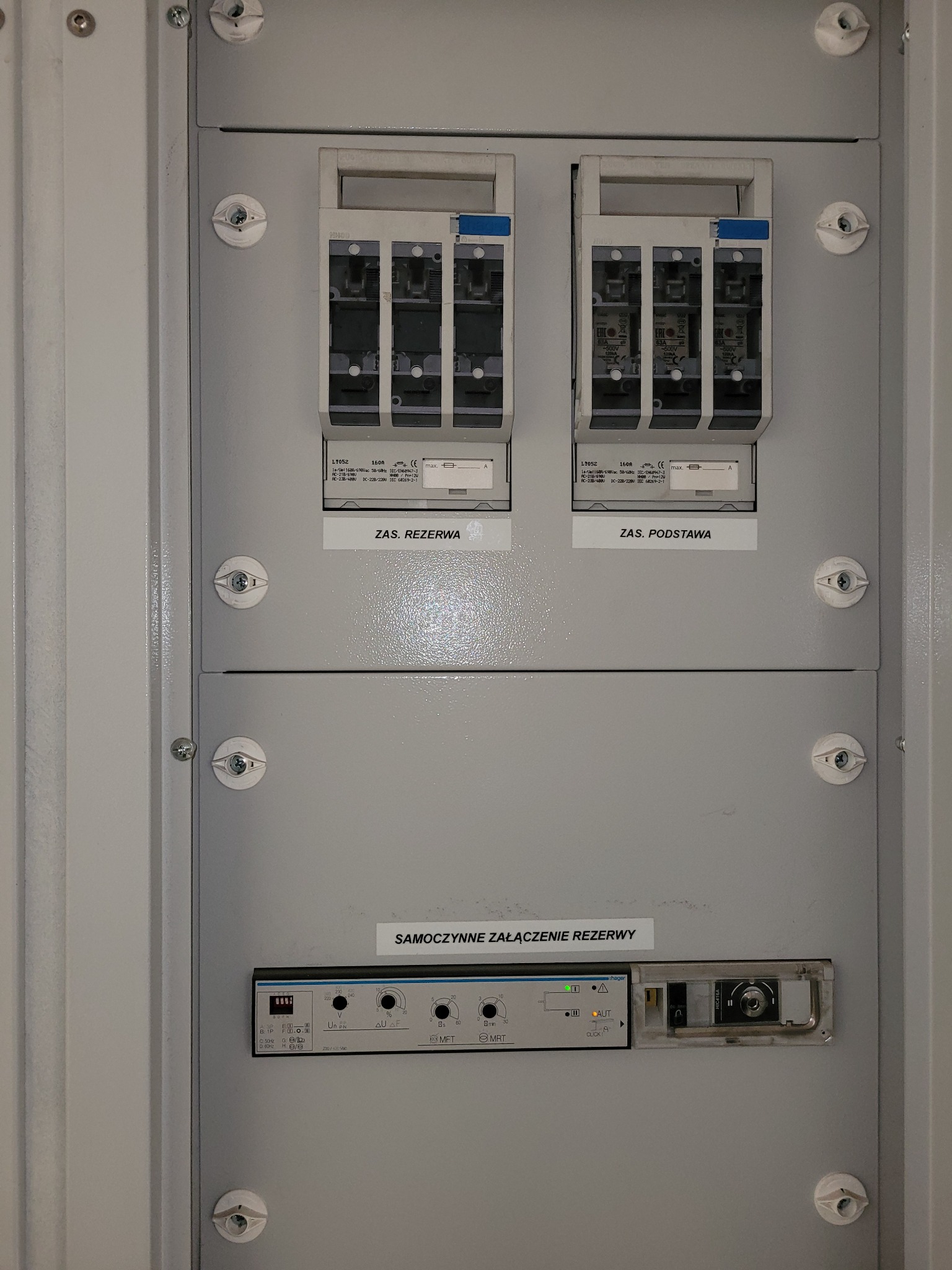 Szara szafa sterownicza z dwoma bezpiecznikami rezerwowymi i podstawowymi oraz modułem automatycznego załączania rezerwy, widoczne napisy i oznaczenia na panelu.