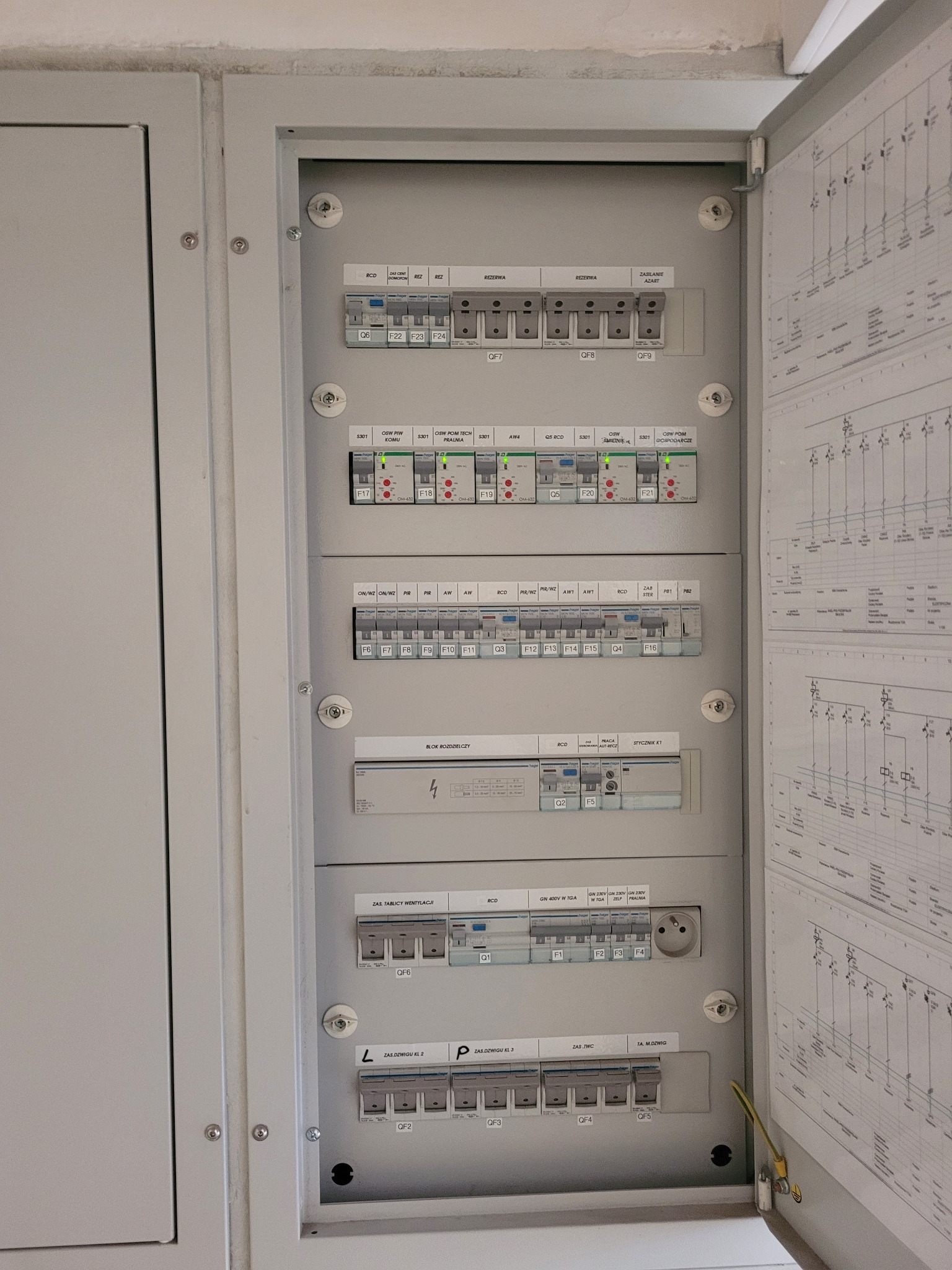 Otwarta skrzynka rozdzielcza z bezpiecznikami i schematami elektrycznymi, widoczne oznaczenia i przełączniki, szara obudowa.